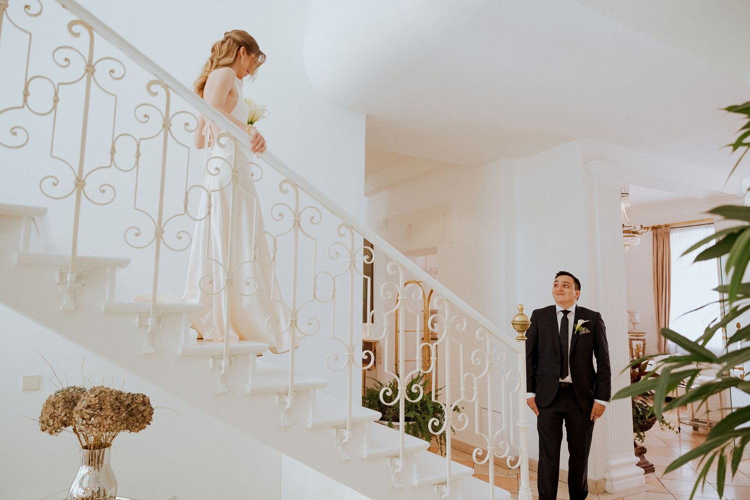 Bride in white gown descends elegant staircase as groom in black suit waits below during wedding first look.
