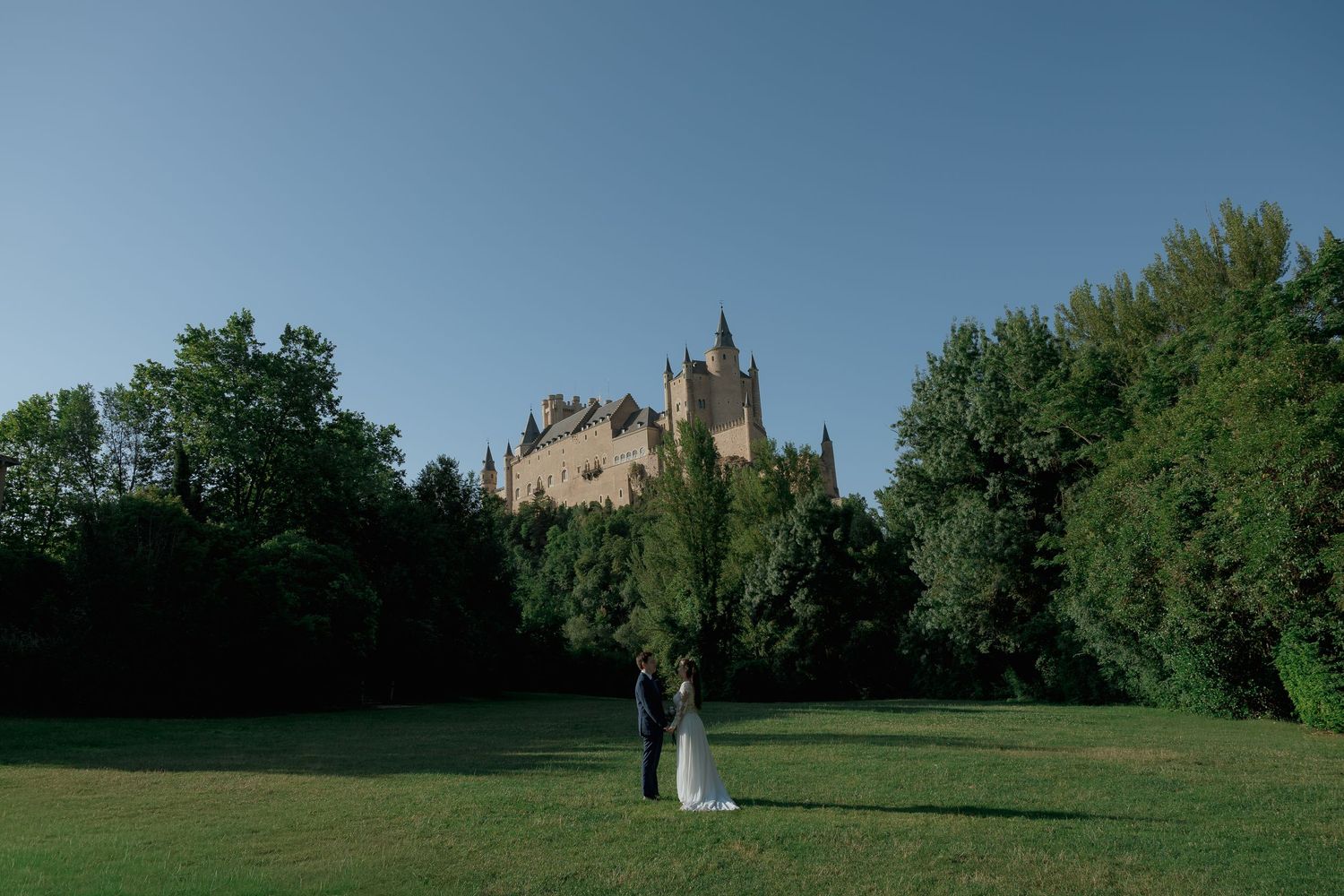Wedding couple stands on green lawn before the historic Alcazar of Segovia castle under clear blue sky.