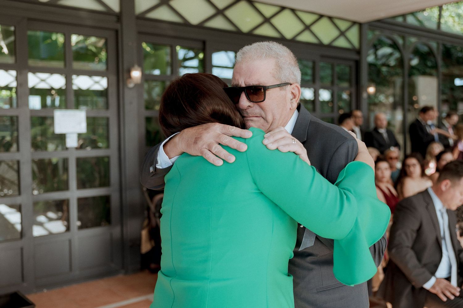 An elderly man in sunglasses shares a warm hug with a woman in a green dress at an outdoor event.