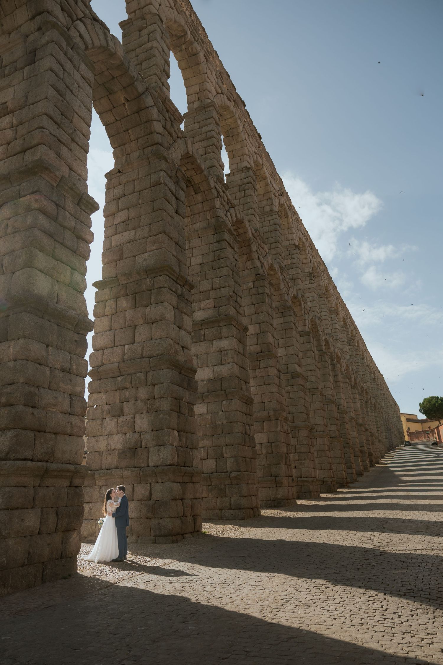 Bride and groom embrace beside the ancient Roman aqueduct of Segovia, Spain, under a blue sky.