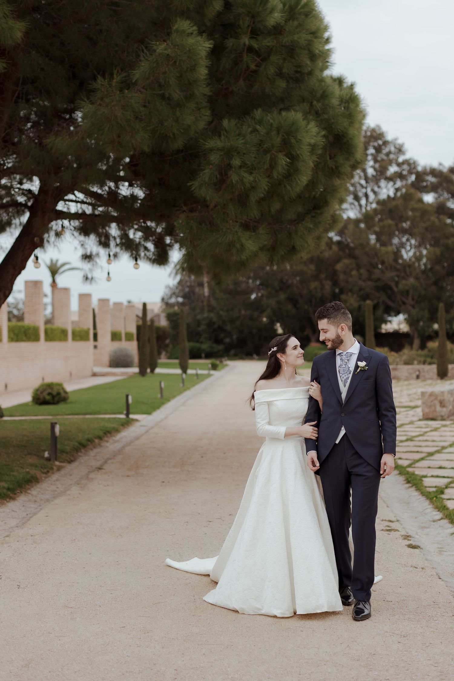 A bride and groom walk hand in hand along a scenic garden path, smiling at each other on their wedding day.
