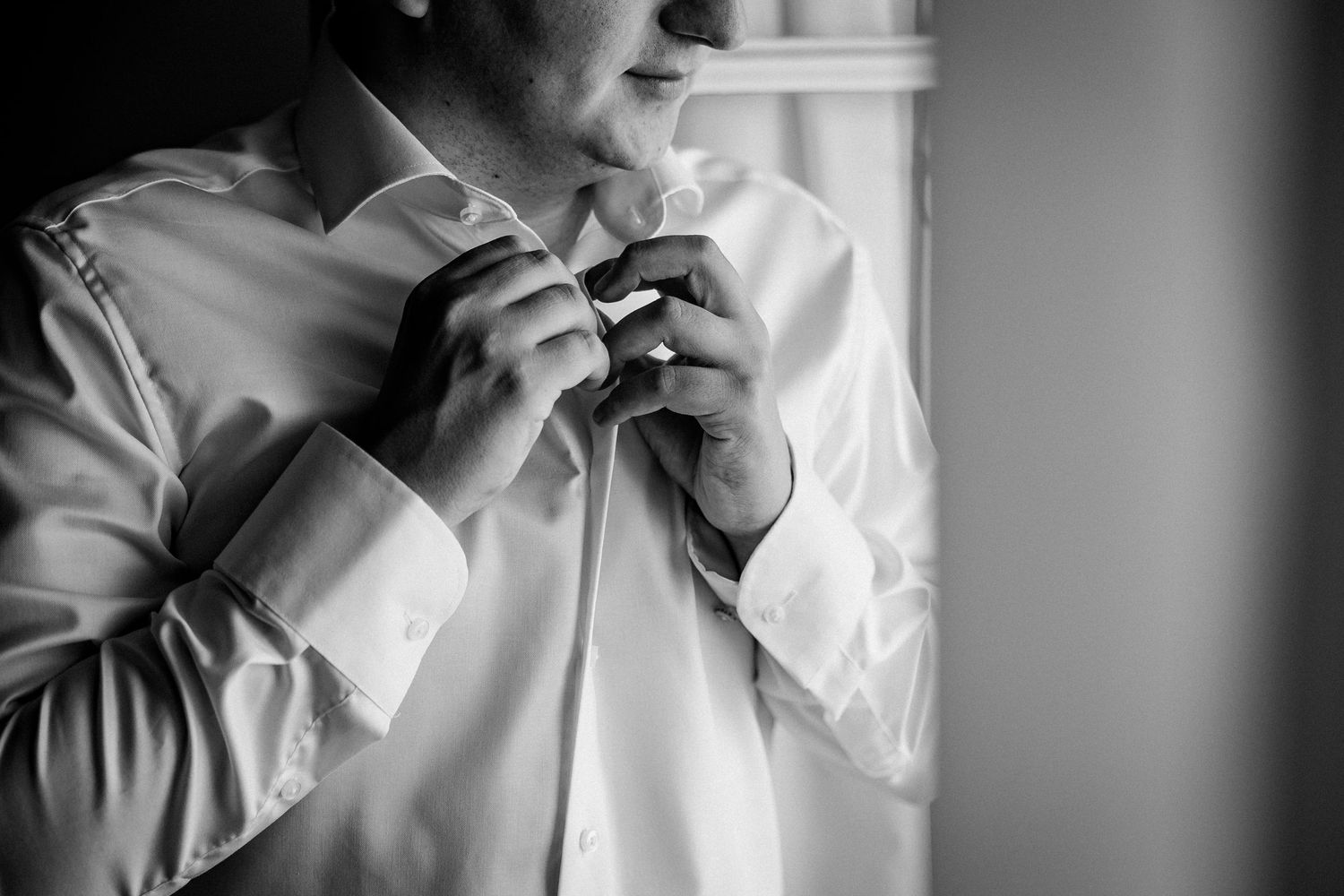 Black and white photo of a groom adjusting his tie while getting dressed in a white dress shirt.