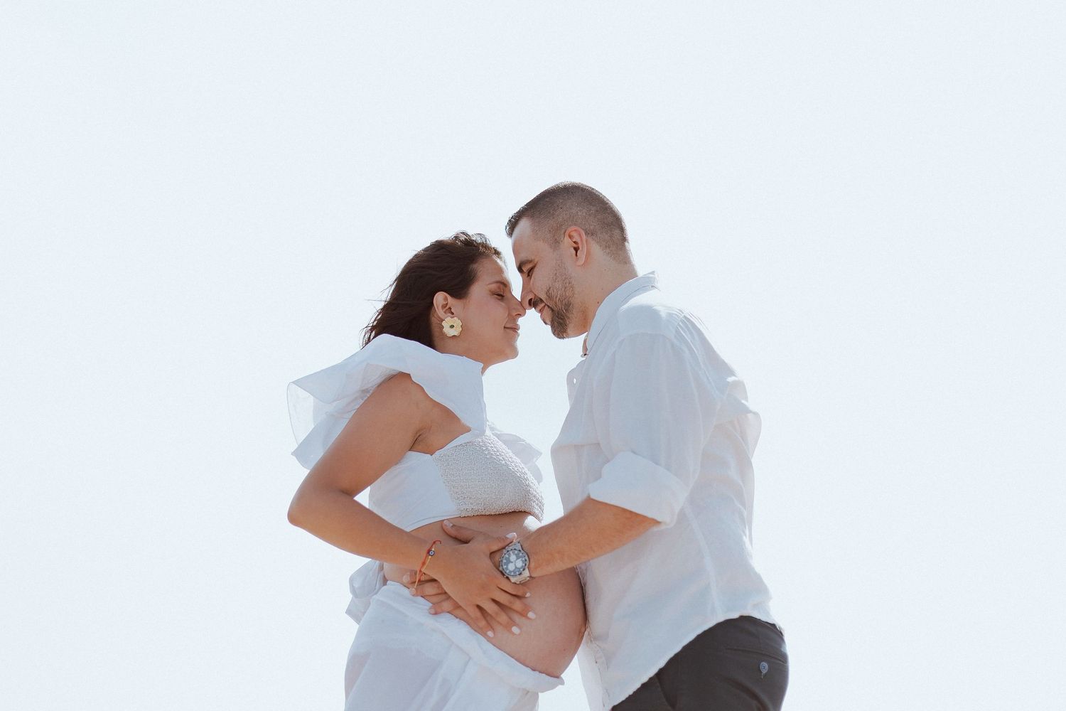A couple in white clothing share an intimate moment against a bright sky background.