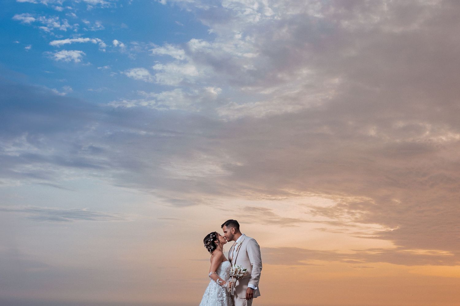 Bride and groom share a romantic kiss against a stunning sunset sky with soft clouds at their outdoor wedding.
