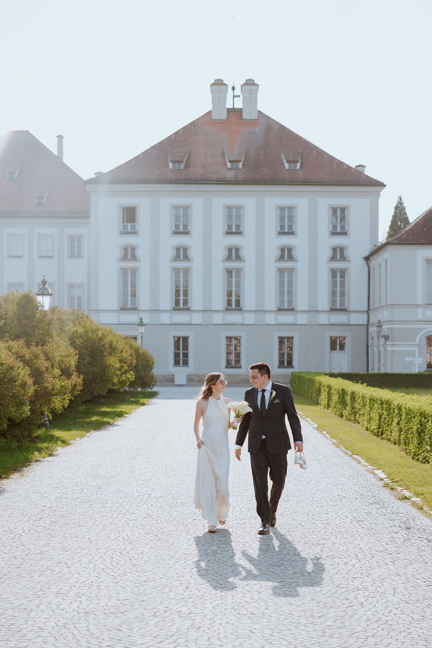 A bride and groom walk hand in hand along a grand cobblestone path in front of a elegant white European palace.