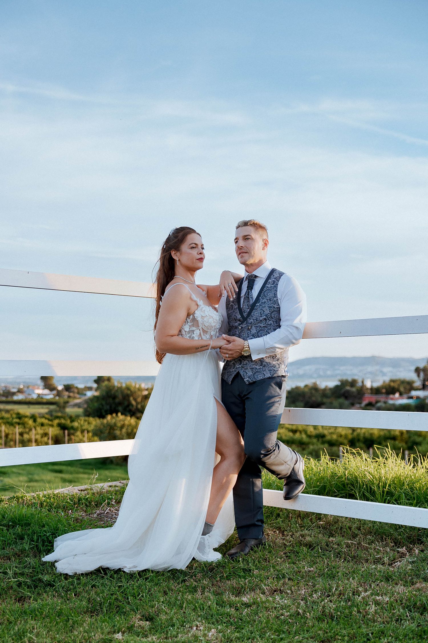 Bride and groom posing by a white fence at a vineyard wedding venue under a blue sky.