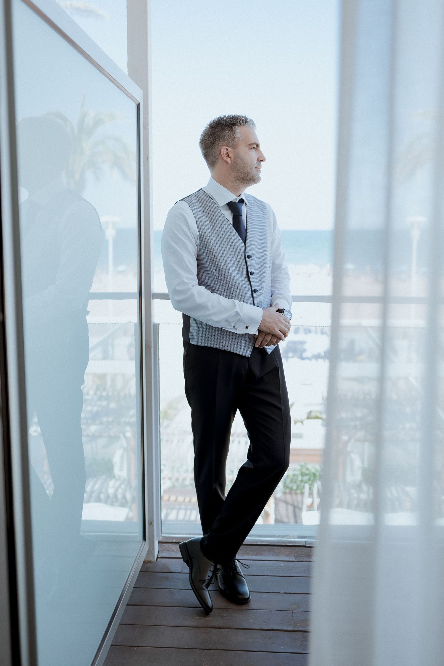 Elegant groom in vest and tie stands by floor-to-ceiling windows overlooking a beach resort.