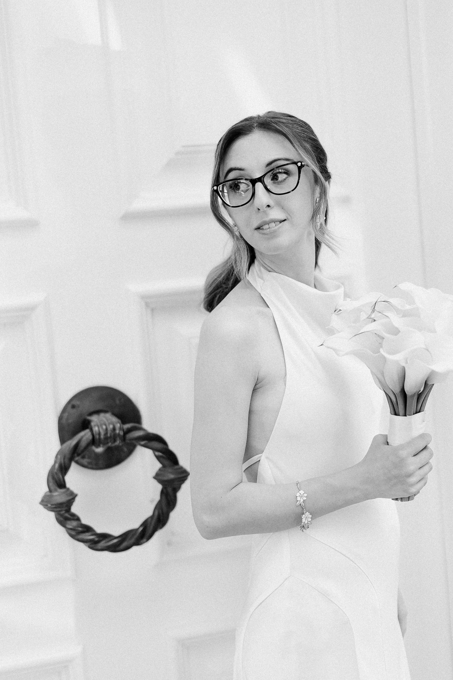 Black and white photo of a bride in a white halter dress holding flowers, wearing glasses, standing by an elegant door.