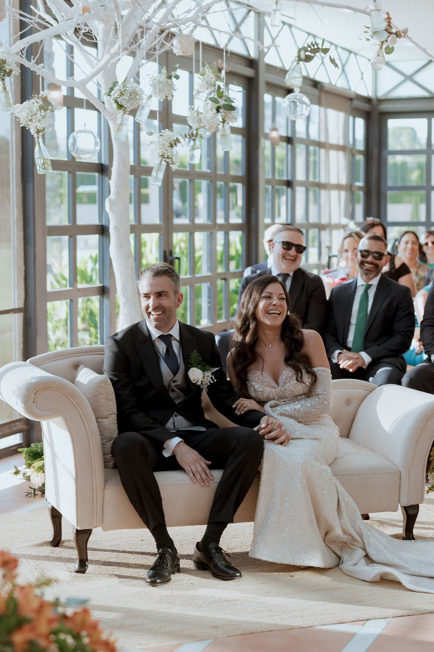 Smiling bride and groom sit together on a white sofa during their elegant indoor wedding ceremony with floral decor.