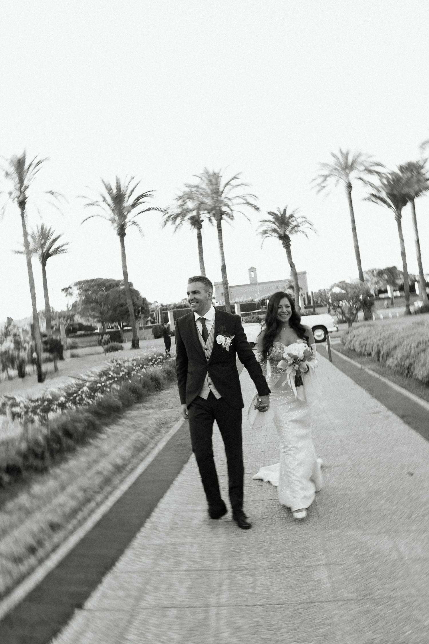 Black and white photo of bride and groom walking hand in hand along a palm tree-lined pathway.
