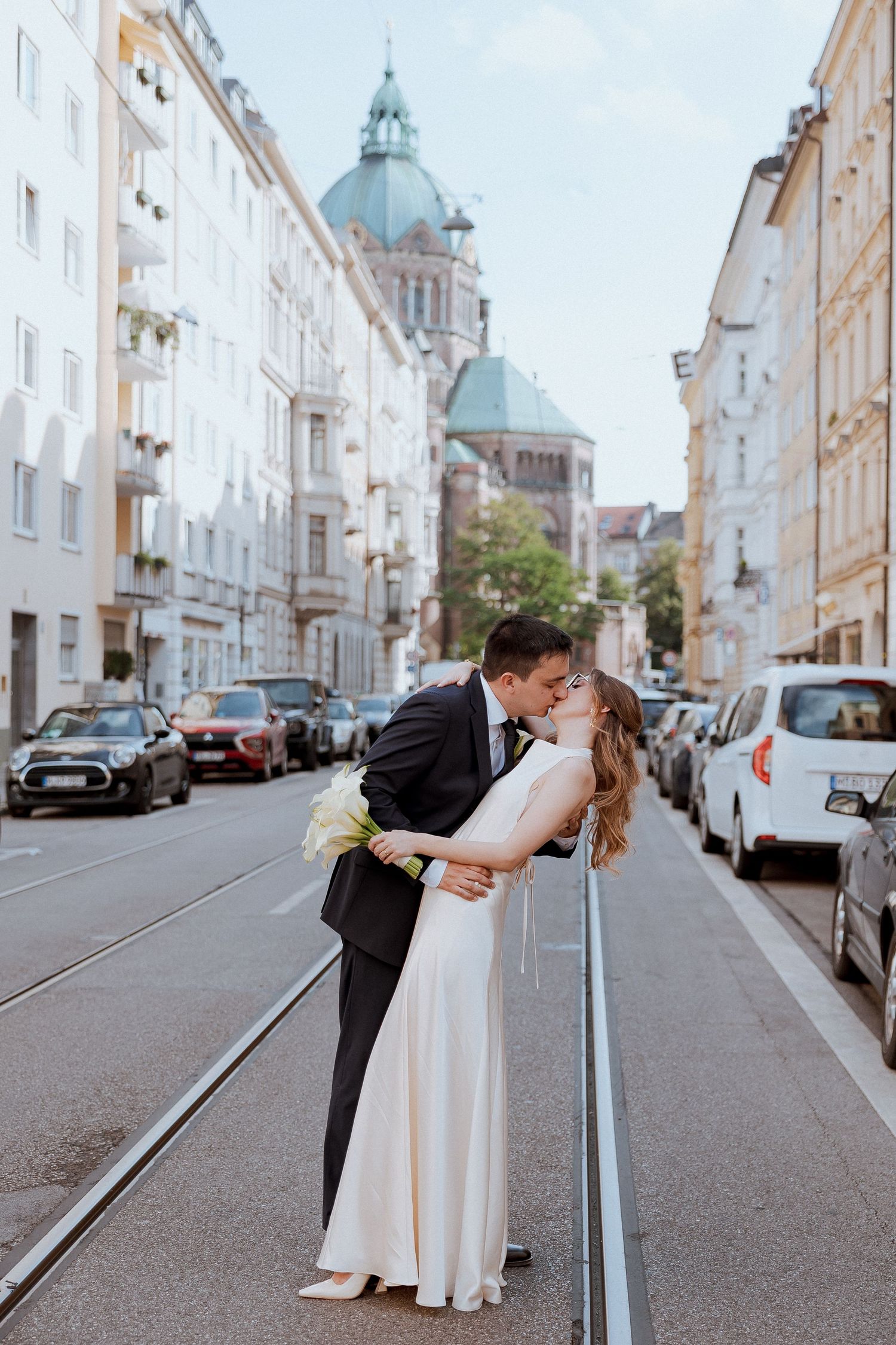 Bride and groom sharing romantic kiss on city tram tracks with European architecture backdrop.