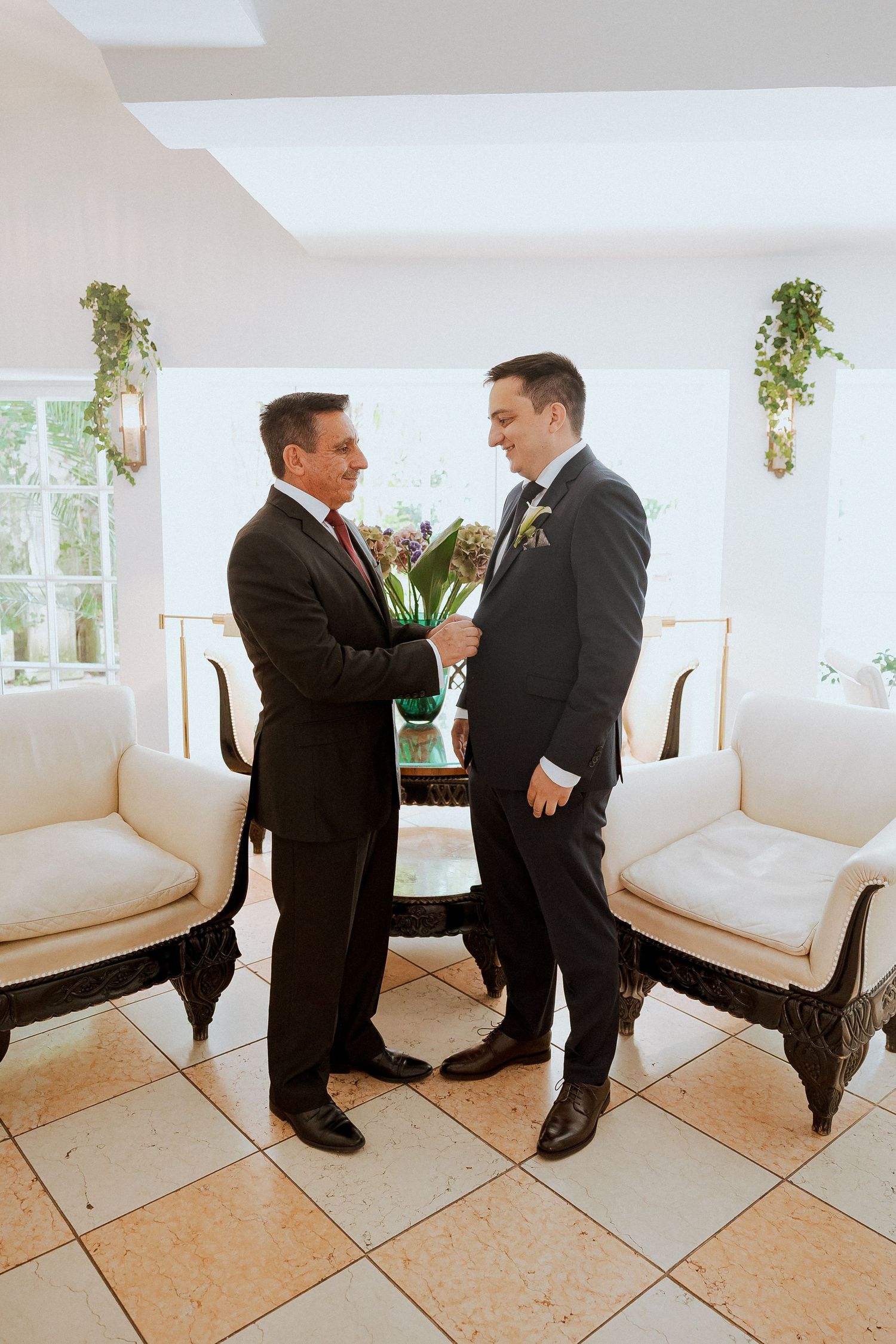 Two men in suits share a warm moment in an elegant white room with ornate chairs during wedding preparations.