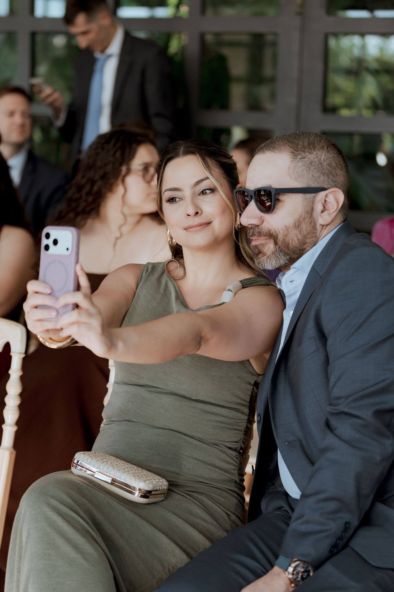 Wedding guests taking a selfie, woman in olive dress and man in sunglasses and suit smiling at phone.