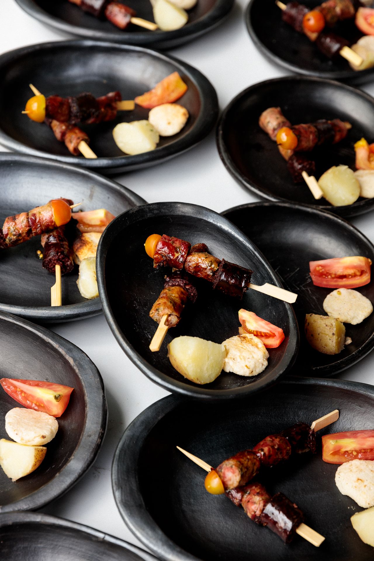 Small black plates arranged in rows featuring grilled meat skewers with potatoes and vegetables served as appetizers.