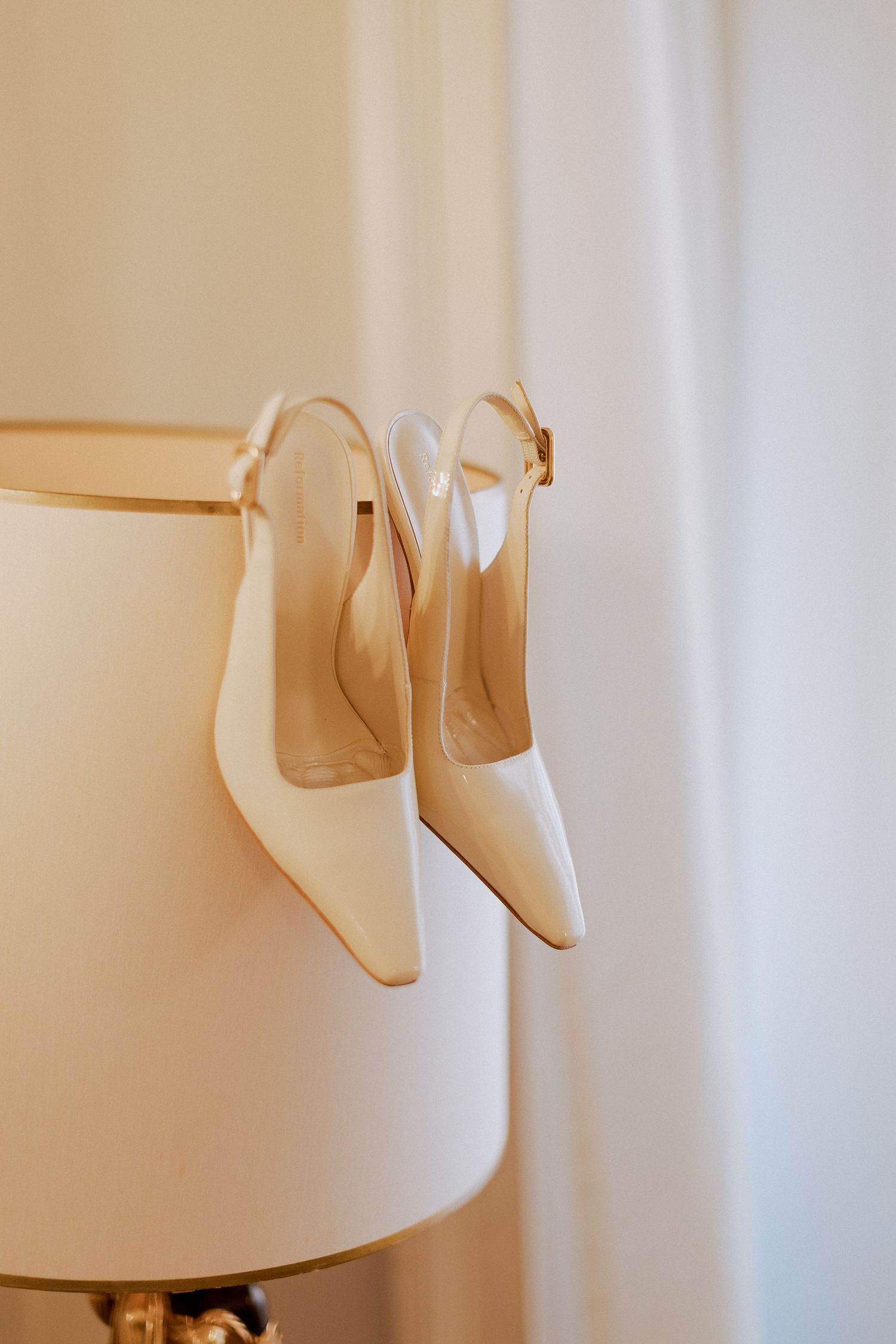Elegant white slingback bridal heels hanging on a cream lampshade before the wedding ceremony.