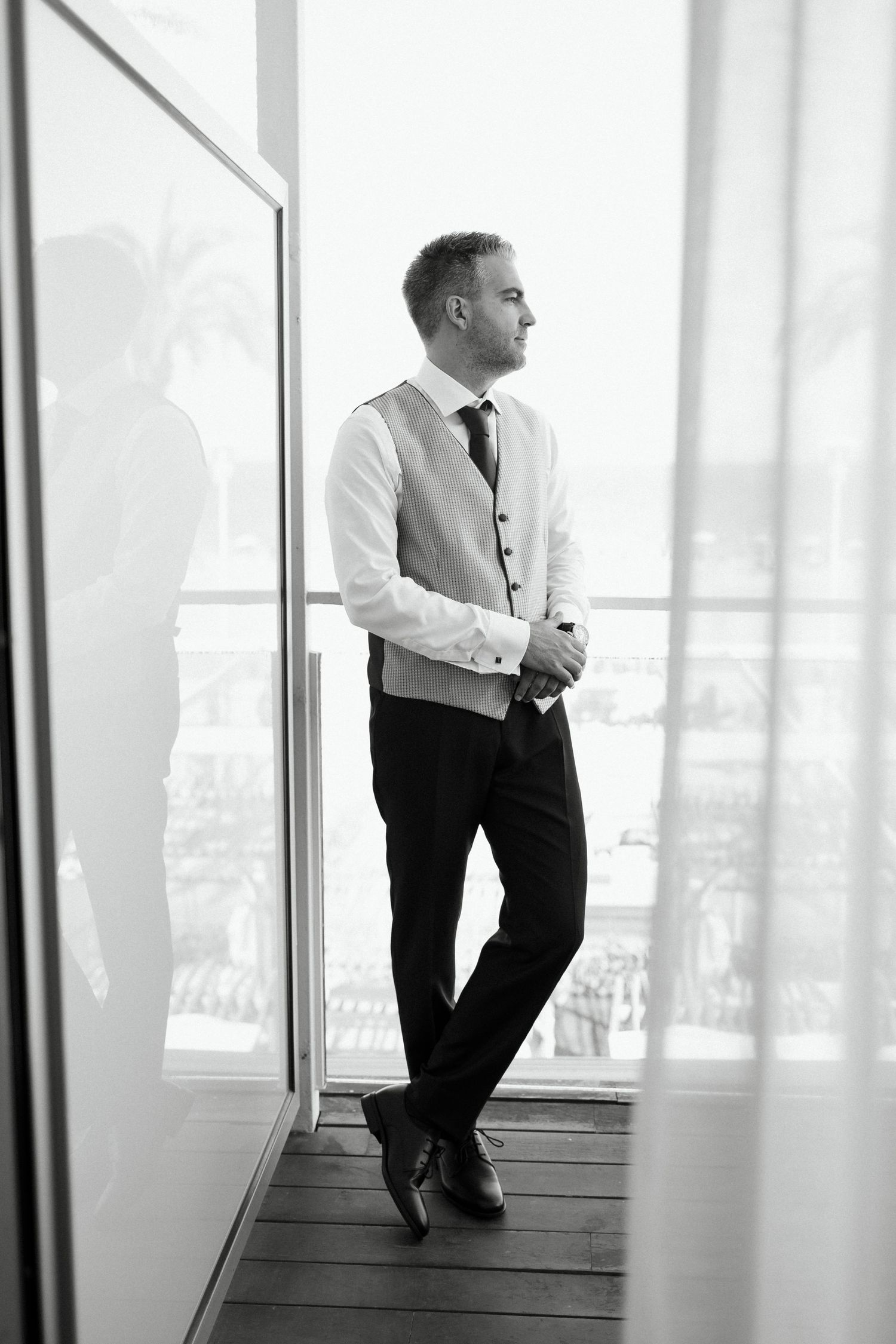 Groom in vest and tie stands by large window in black and white wedding photo.
