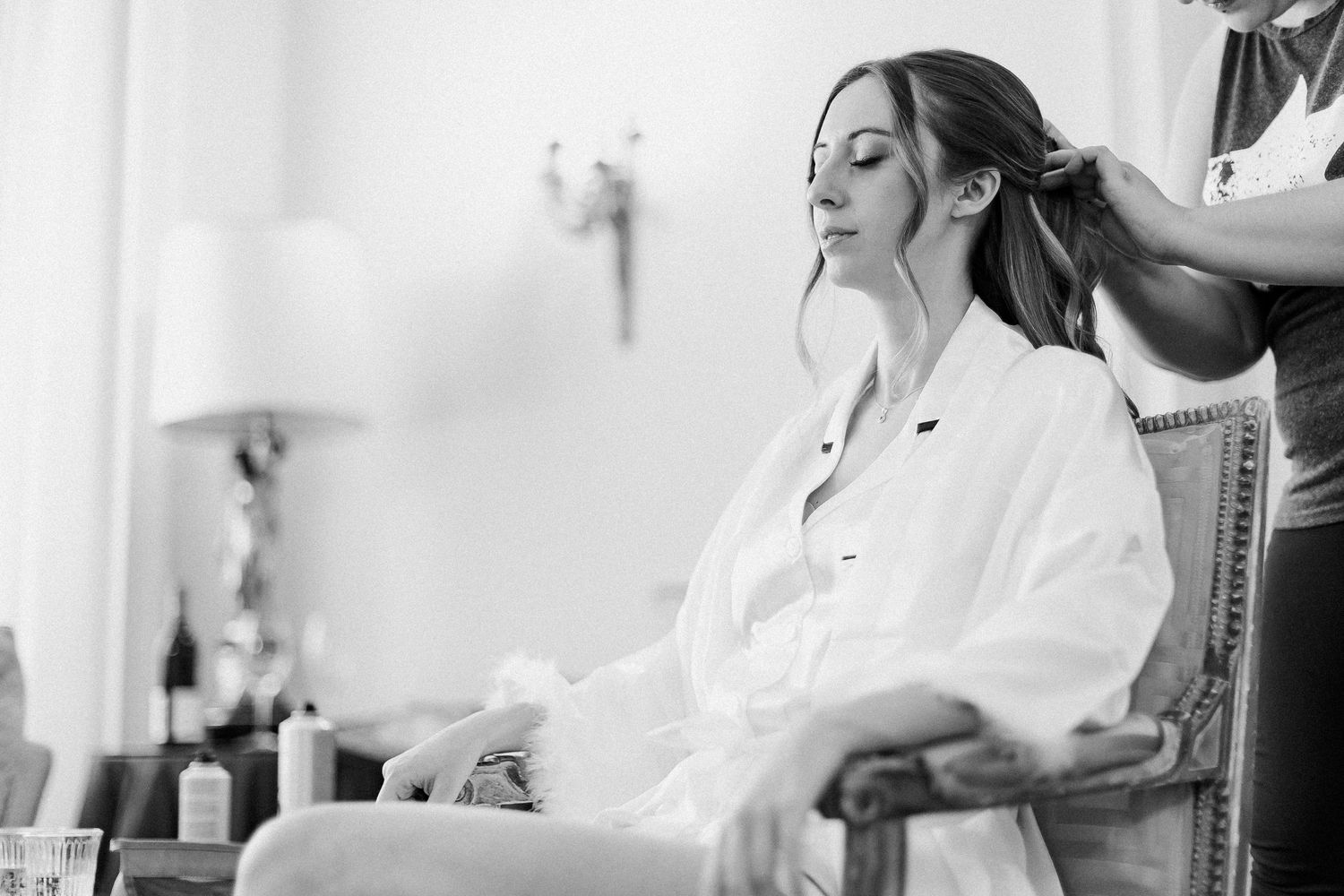 Bride getting hair styled while sitting in chair wearing white robe before wedding ceremony.