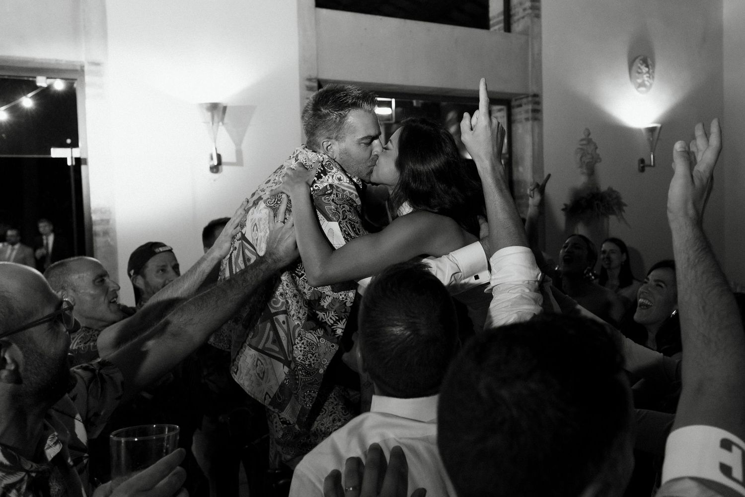 Couple shares romantic kiss on dance floor surrounded by cheering guests at wedding reception.