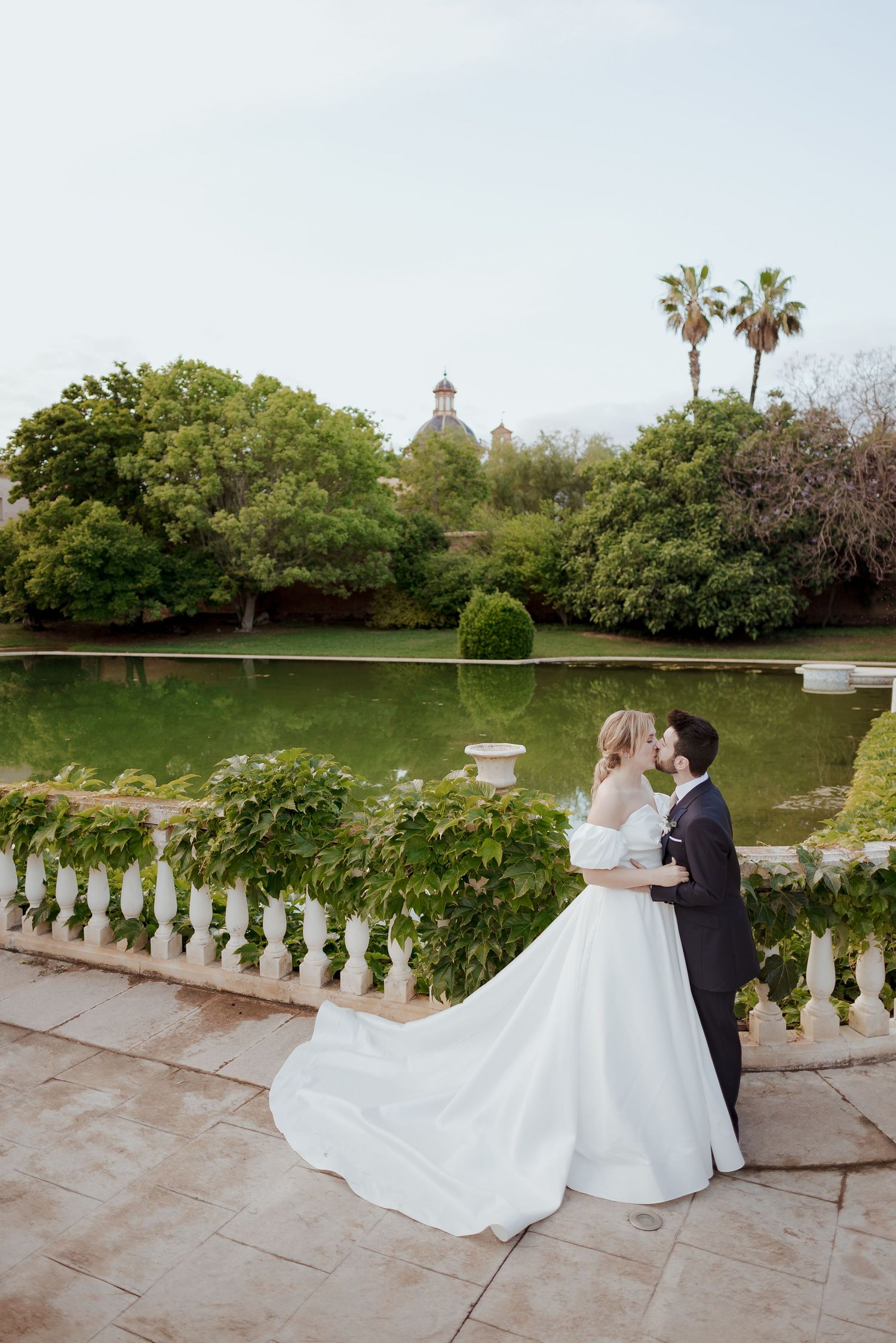 Bride and groom share a romantic kiss on an elegant balustrade terrace surrounded by lush gardens and a serene pond.