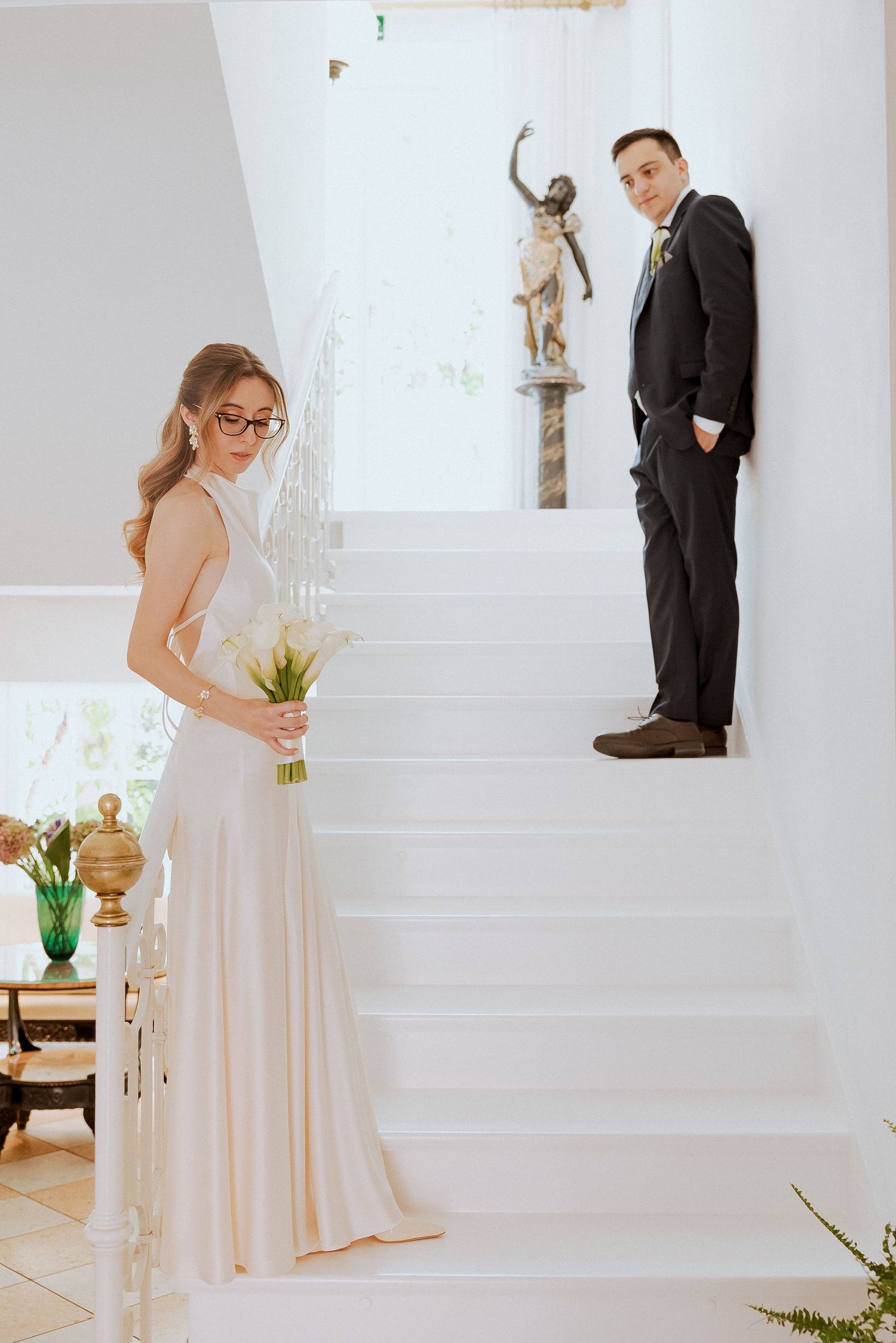 Bride in white gown holding calla lily bouquet stands at staircase base as groom leans on steps above.