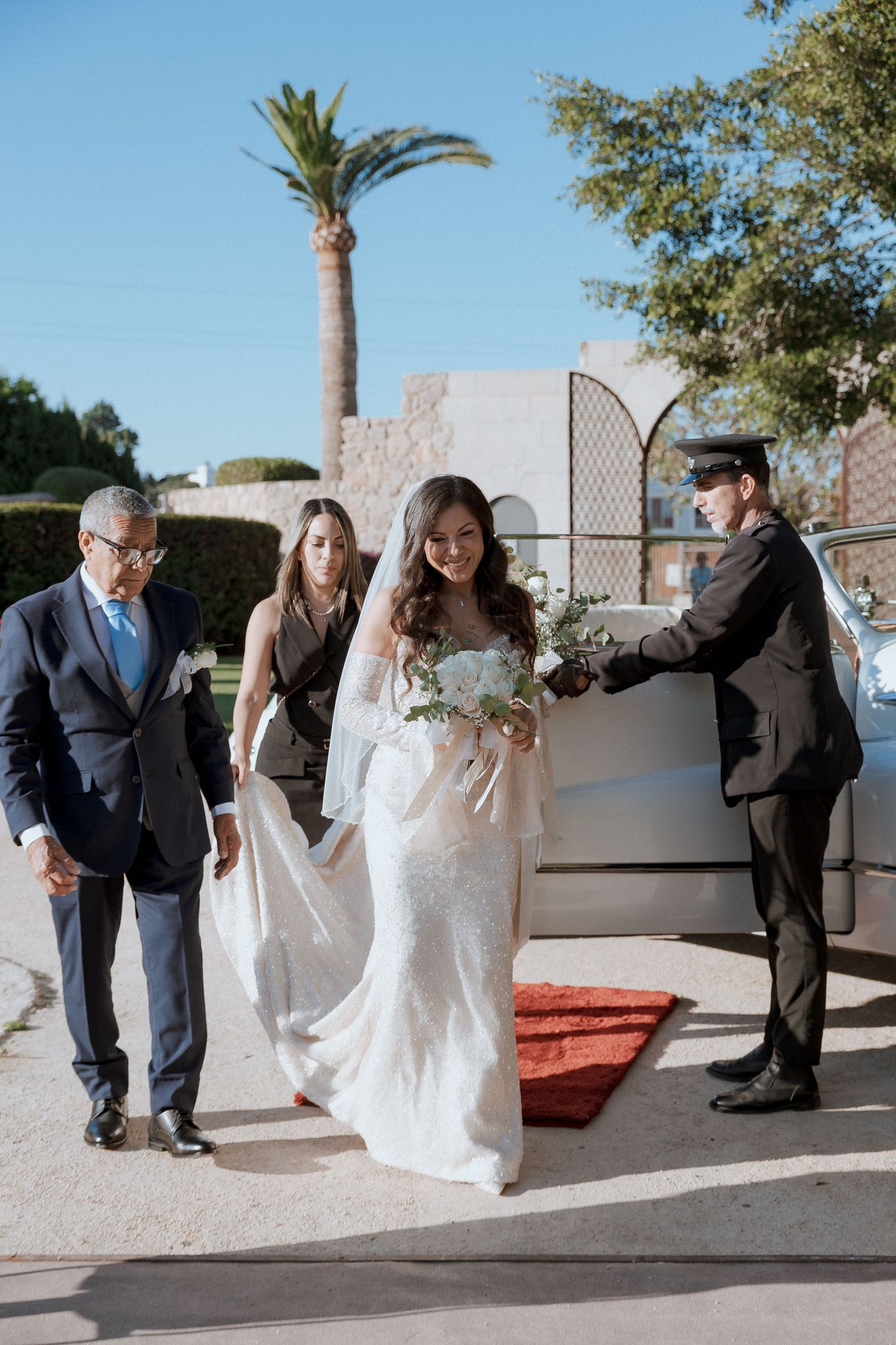 Bride in lace wedding gown arriving with bouquet, assisted by guests near a vintage car outdoors.