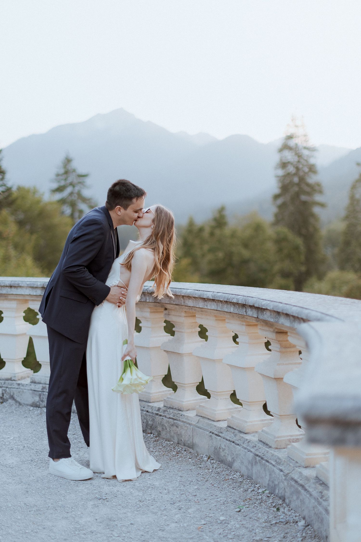 Romantic wedding couple shares an intimate kiss on a stone balcony with mountain views in the background.