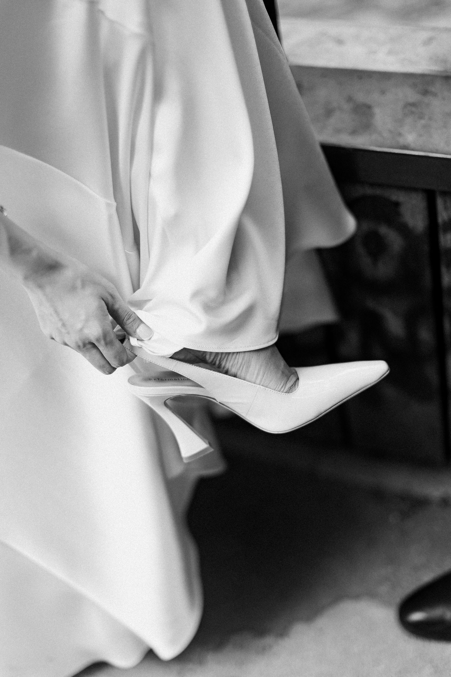 Black and white photo of a bride adjusting her white stiletto heel wedding shoe beneath her flowing bridal gown.