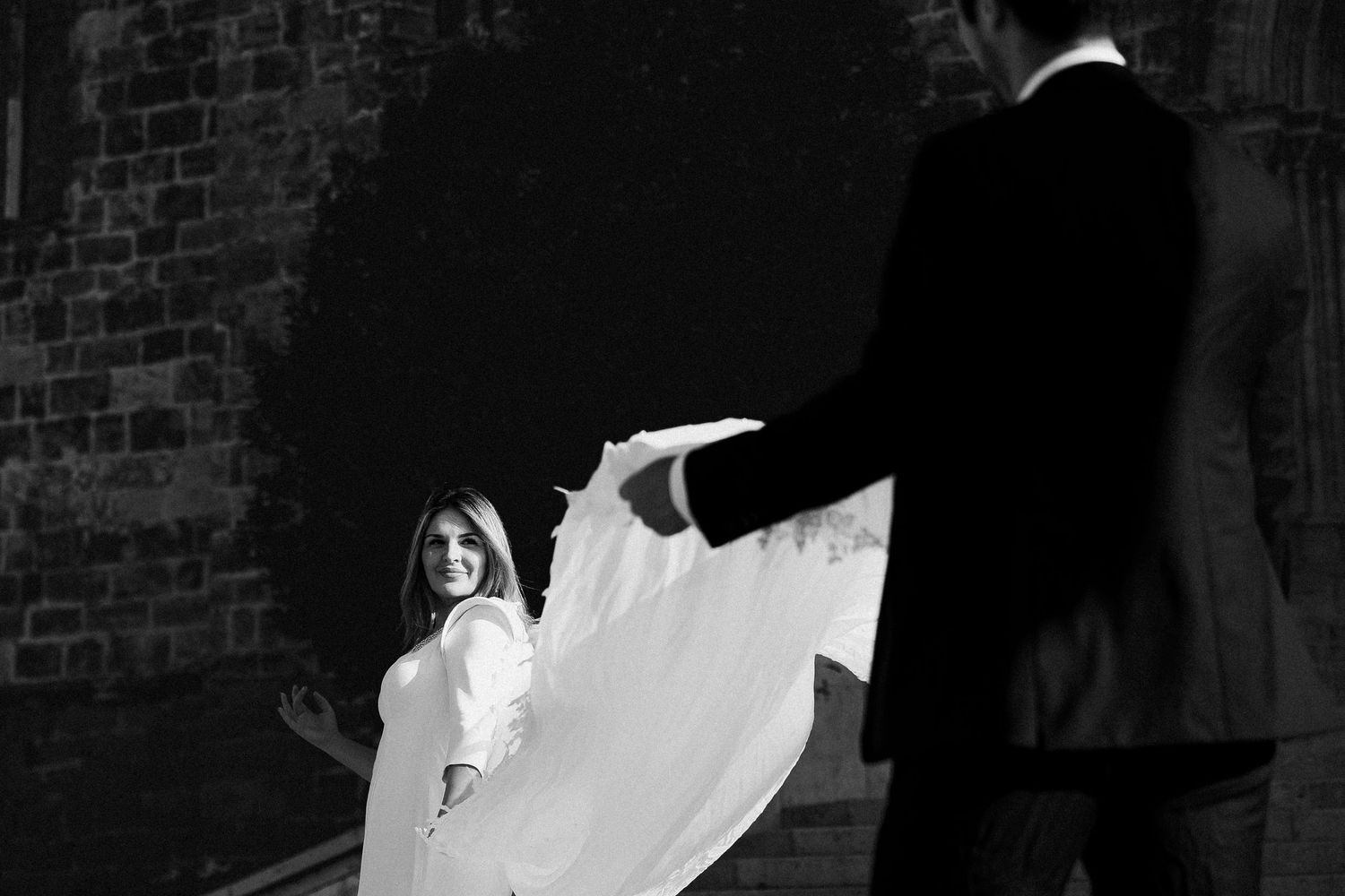 Dramatic black and white wedding photo showing bride in flowing white dress being dipped in front of brick wall.