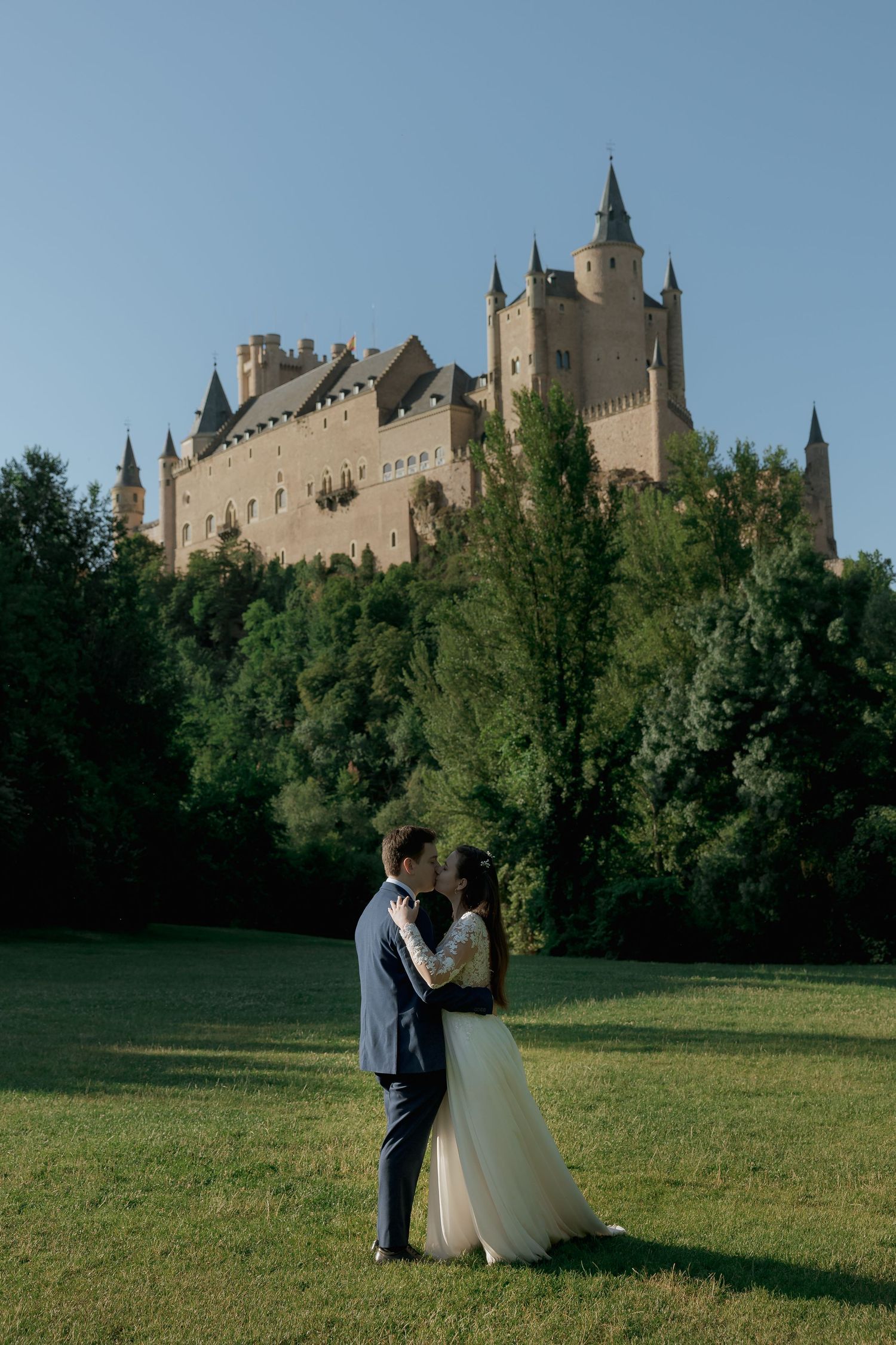 Bride and groom embrace on green lawn with stunning Alcazar of Segovia castle rising above lush trees under clear blue sky.