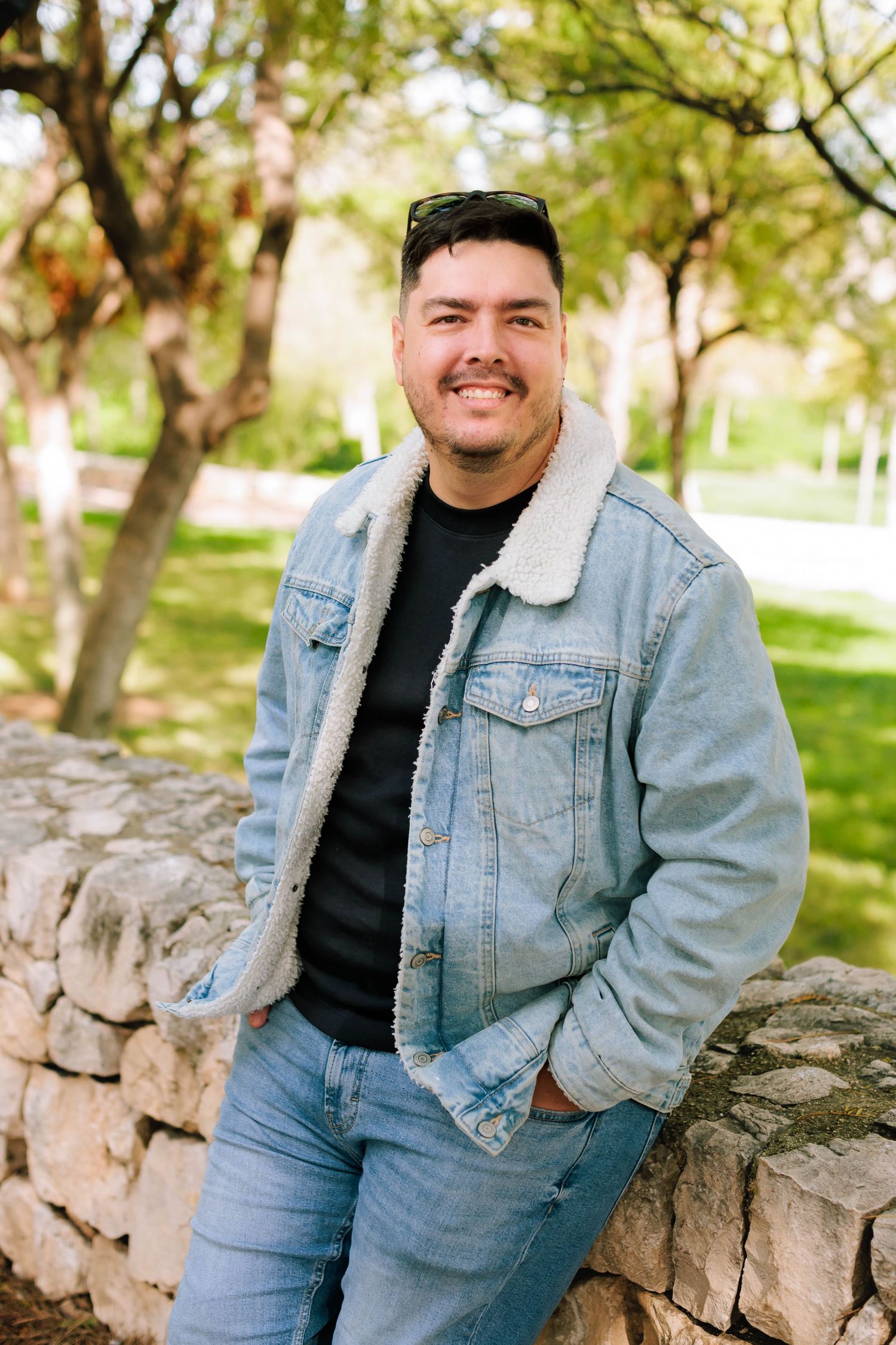 A person in a light denim jacket and black shirt sits on a stone wall in a park setting during spring.