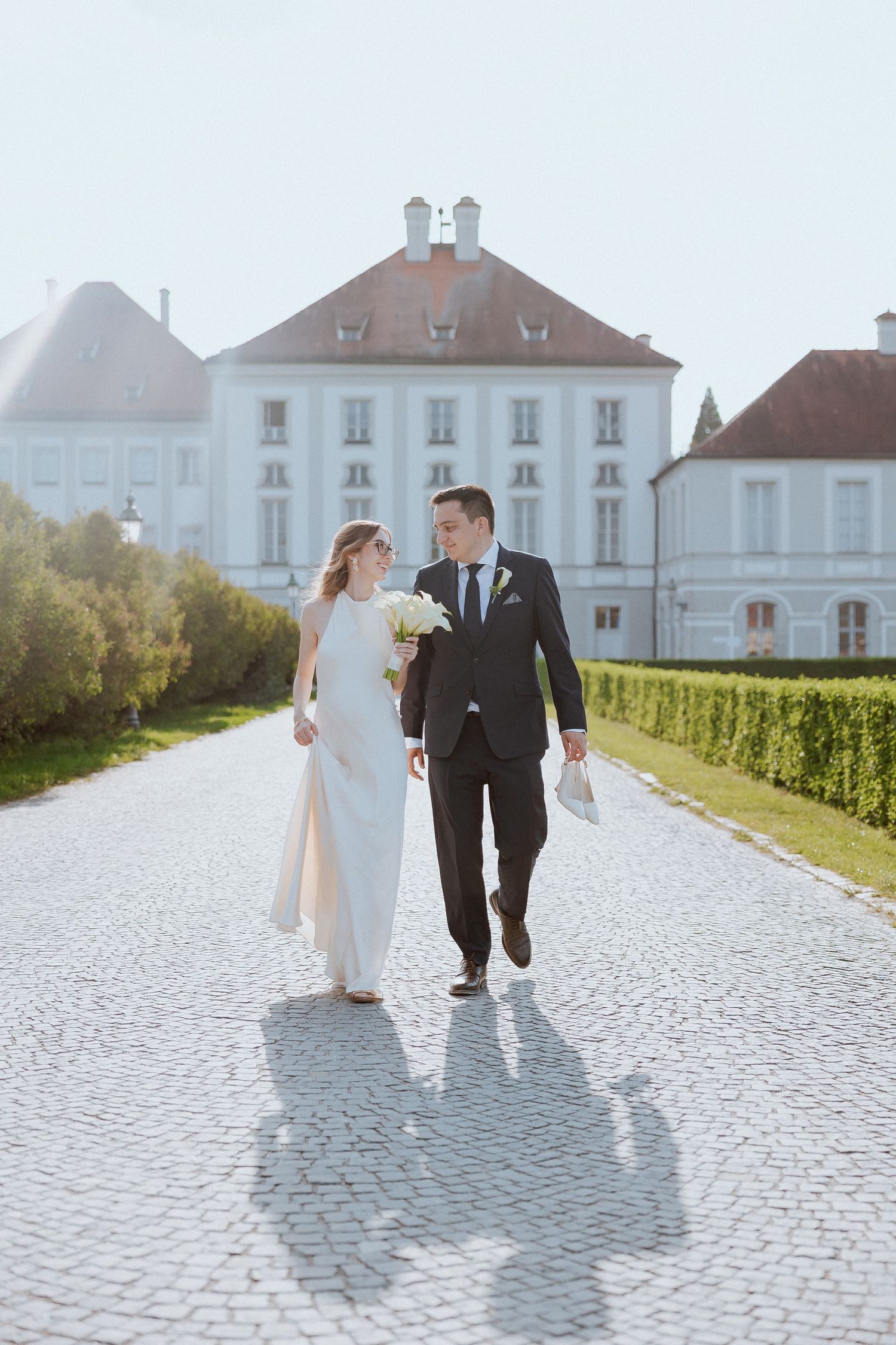 A couple in wedding attire walks together on a cobblestone path in front of elegant white buildings on a sunny day.