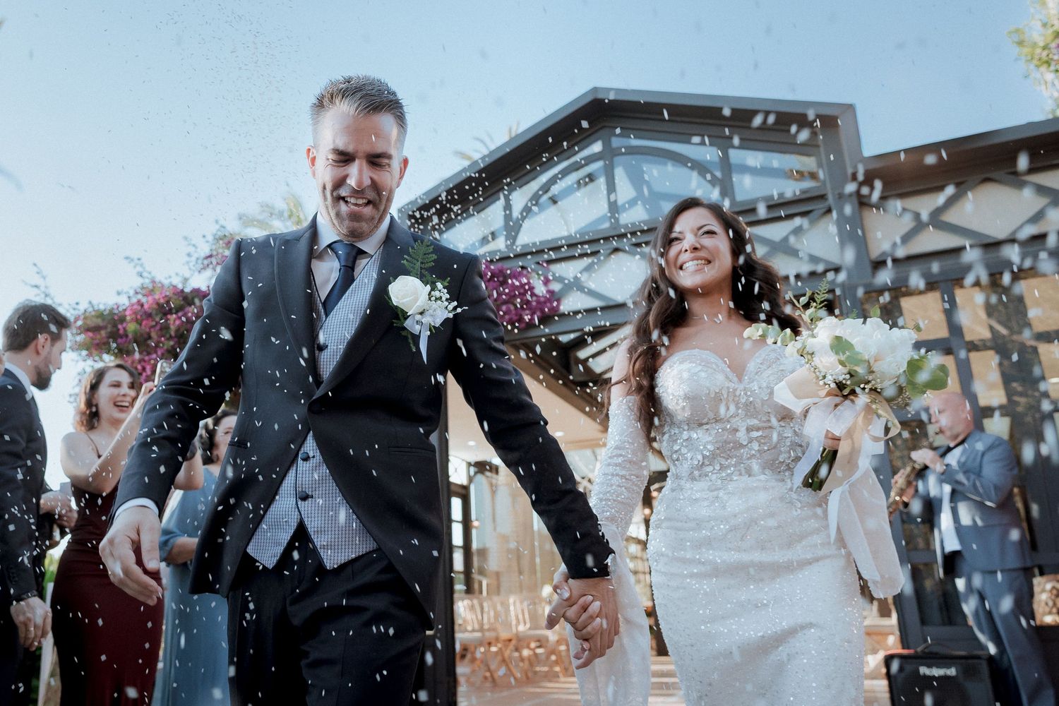Happy bride and groom walking hand in hand through confetti shower outside elegant wedding venue.