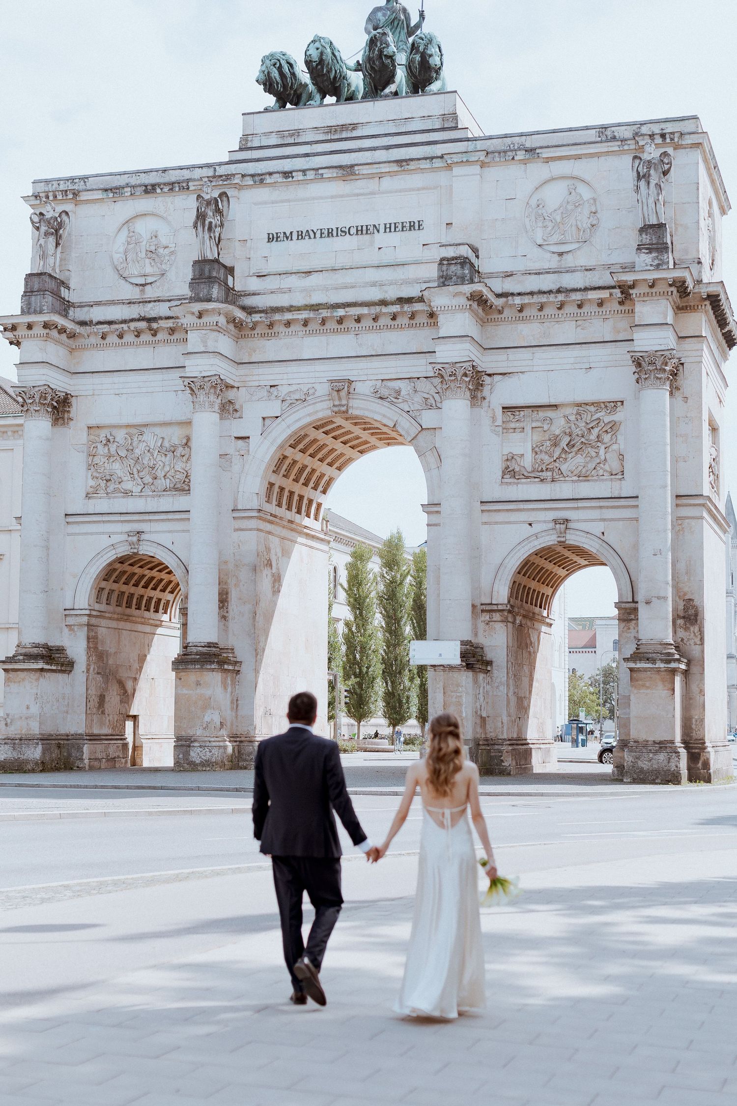 Bride and groom walk hand in hand toward historic triumphal arch in romantic outdoor wedding portrait.