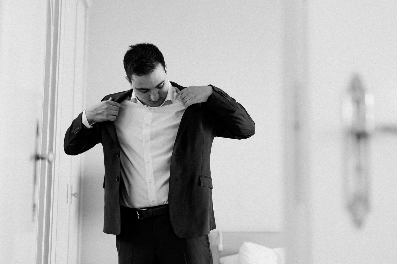 A well-dressed man in a suit adjusting his jacket collar in a bright room, captured in black and white.