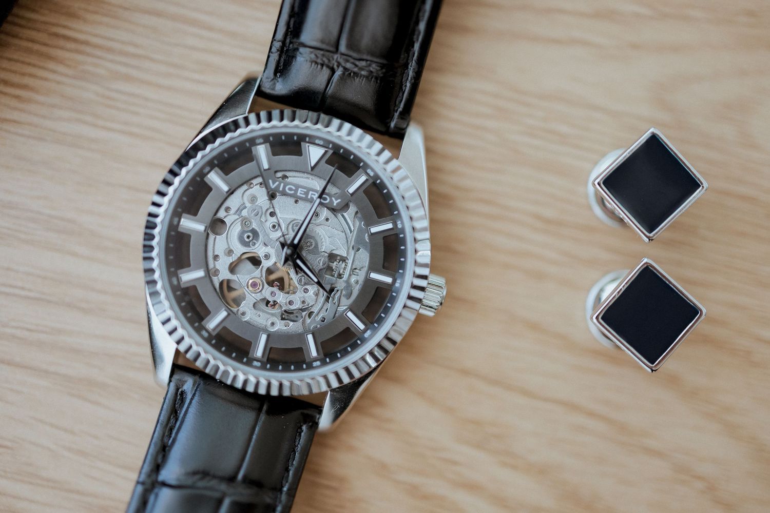 Skeleton mechanical watch with silver case and black leather strap beside two square black and silver cufflinks on wood surface.