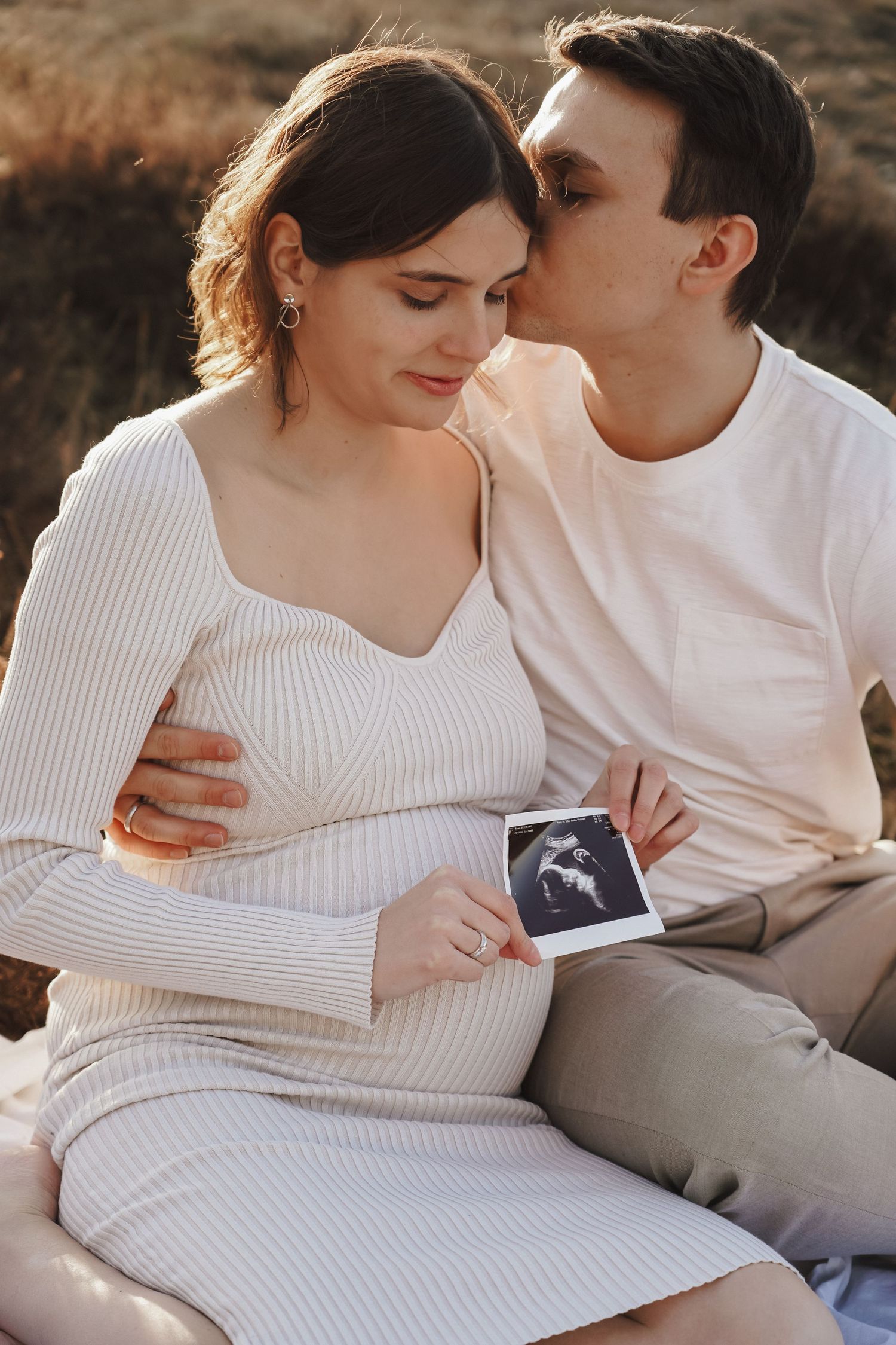 Couple sharing tender moment outdoors while holding ultrasound photo during pregnancy photoshoot.