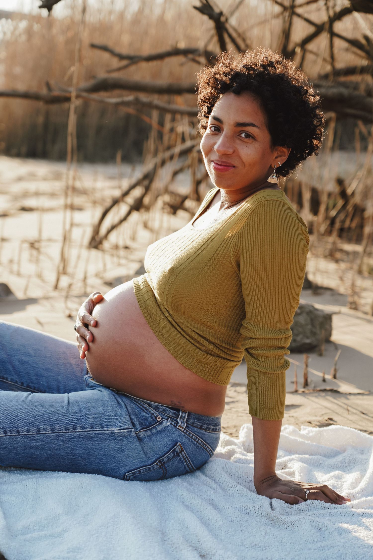 Casual maternity portrait with mustard sweater and jeans on white blanket outdoors.