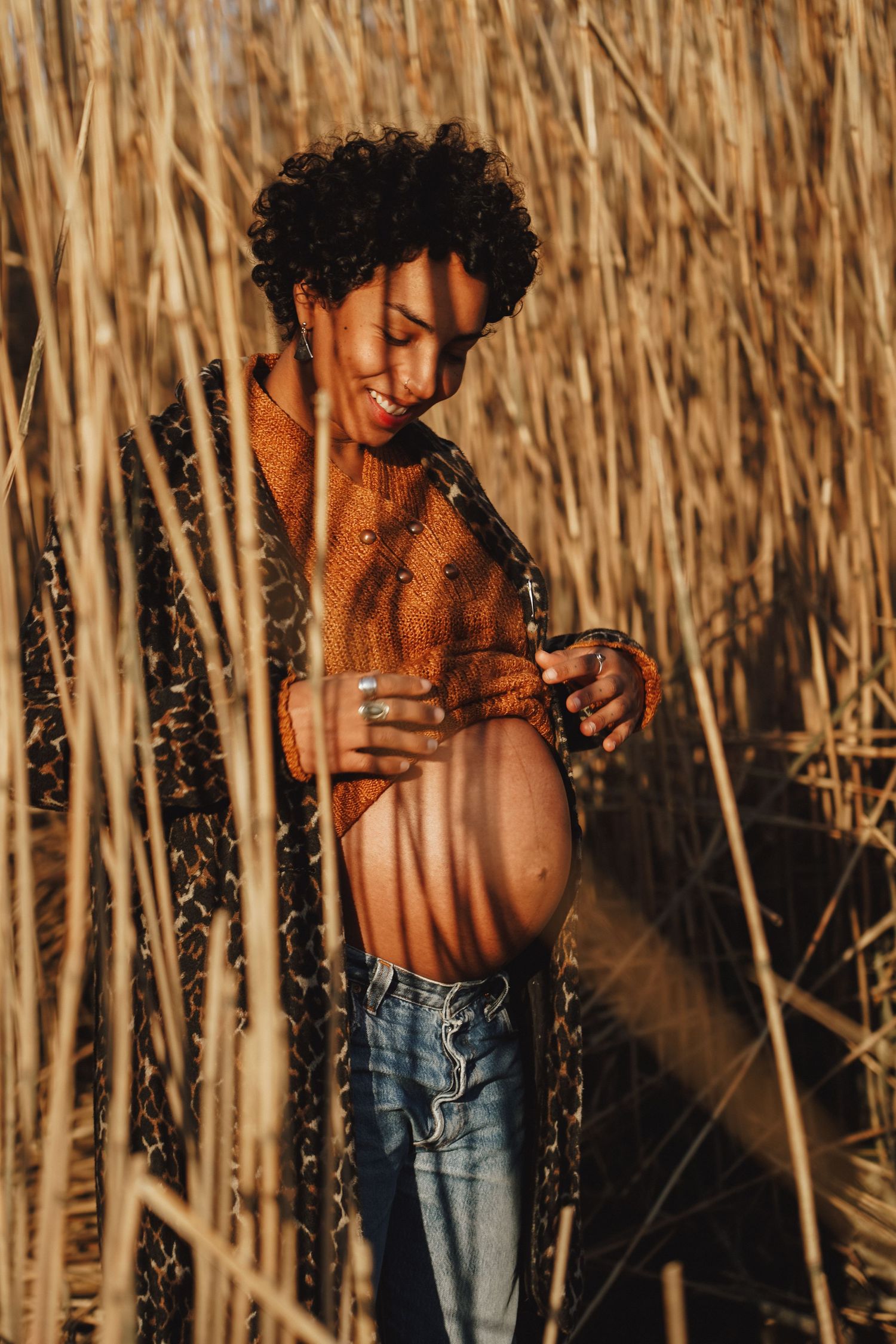Artistic maternity shot through dried reeds with warm sunset lighting.