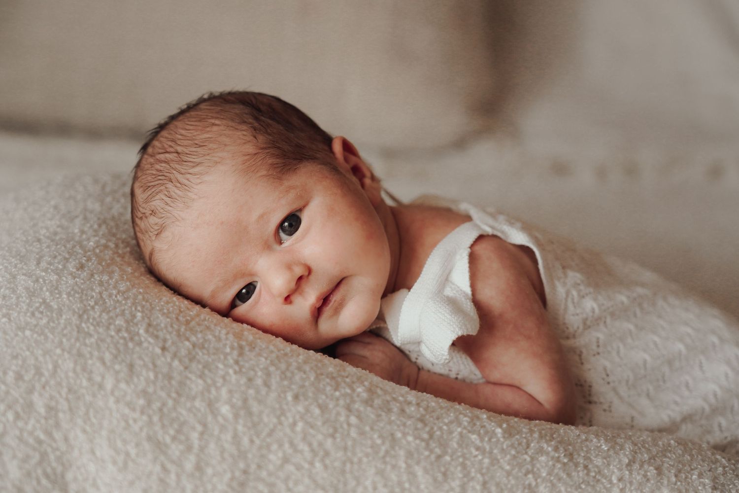 Close up portrait of a newborn baby with bright eyes lying on soft beige fabric.
