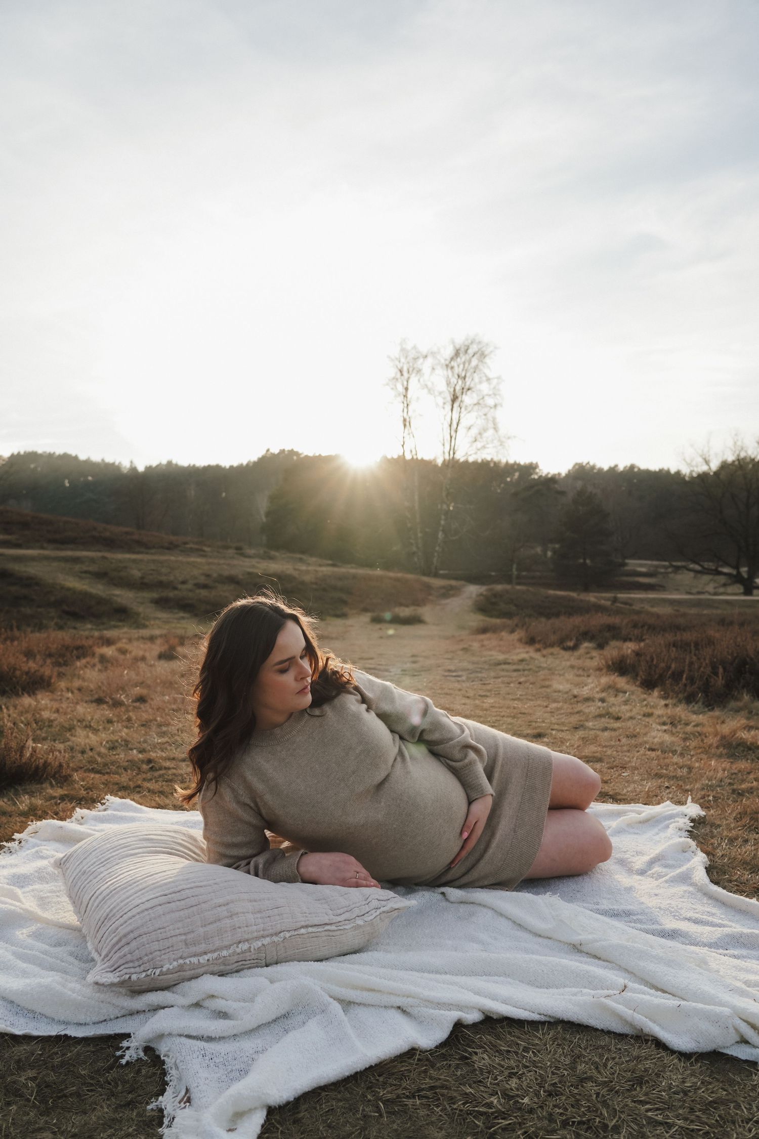 A maternity photo session at sunset with the subject lounging on a white blanket in a grey sweater dress.