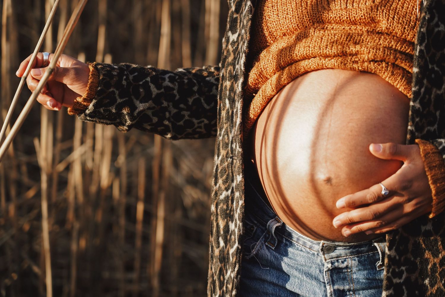Close-up shot of a pregnant belly with hands resting on it against a backdrop of autumn foliage.