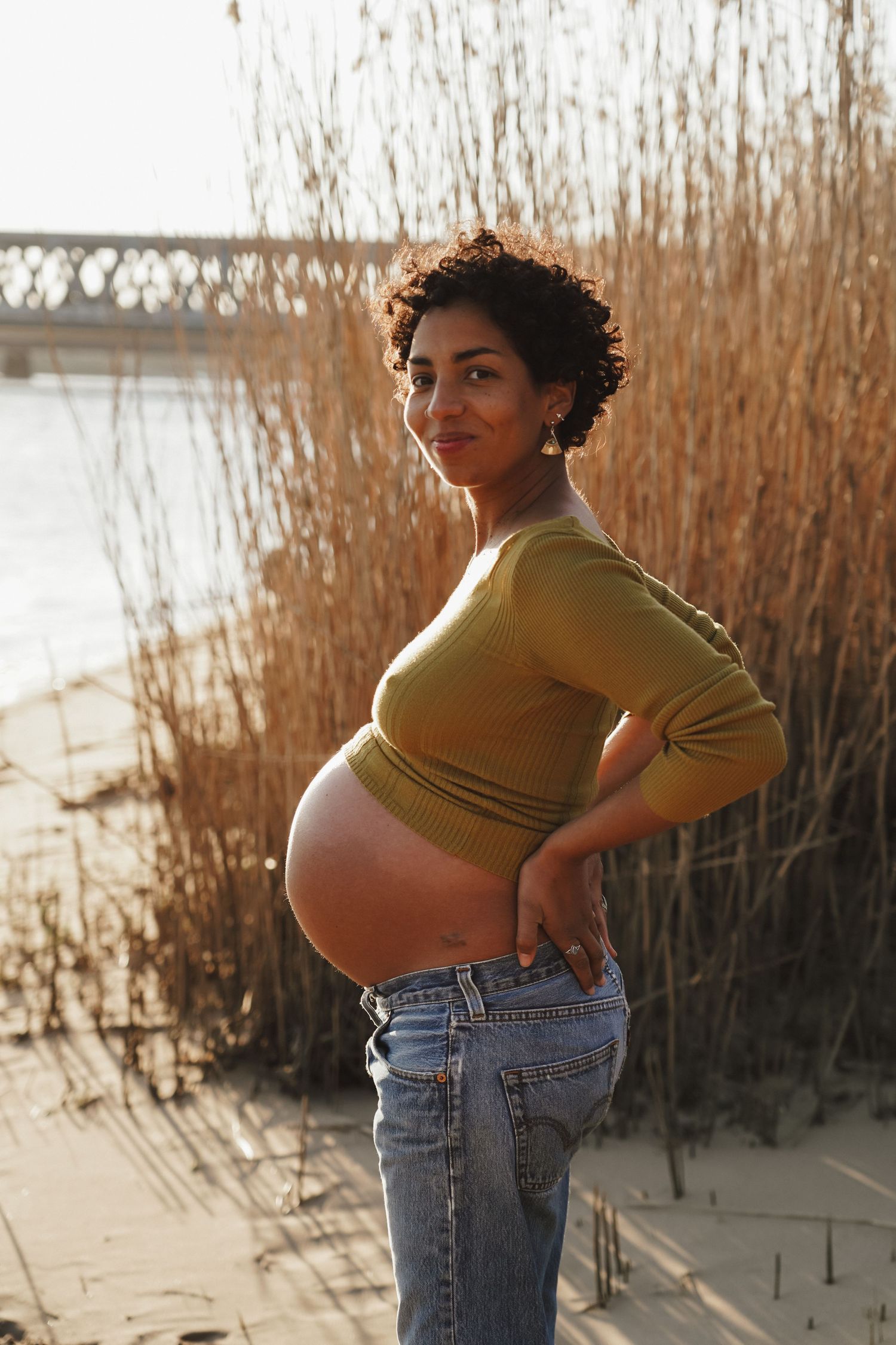 Side profile maternity portrait against golden reeds at sunset wearing yellow top and jeans.