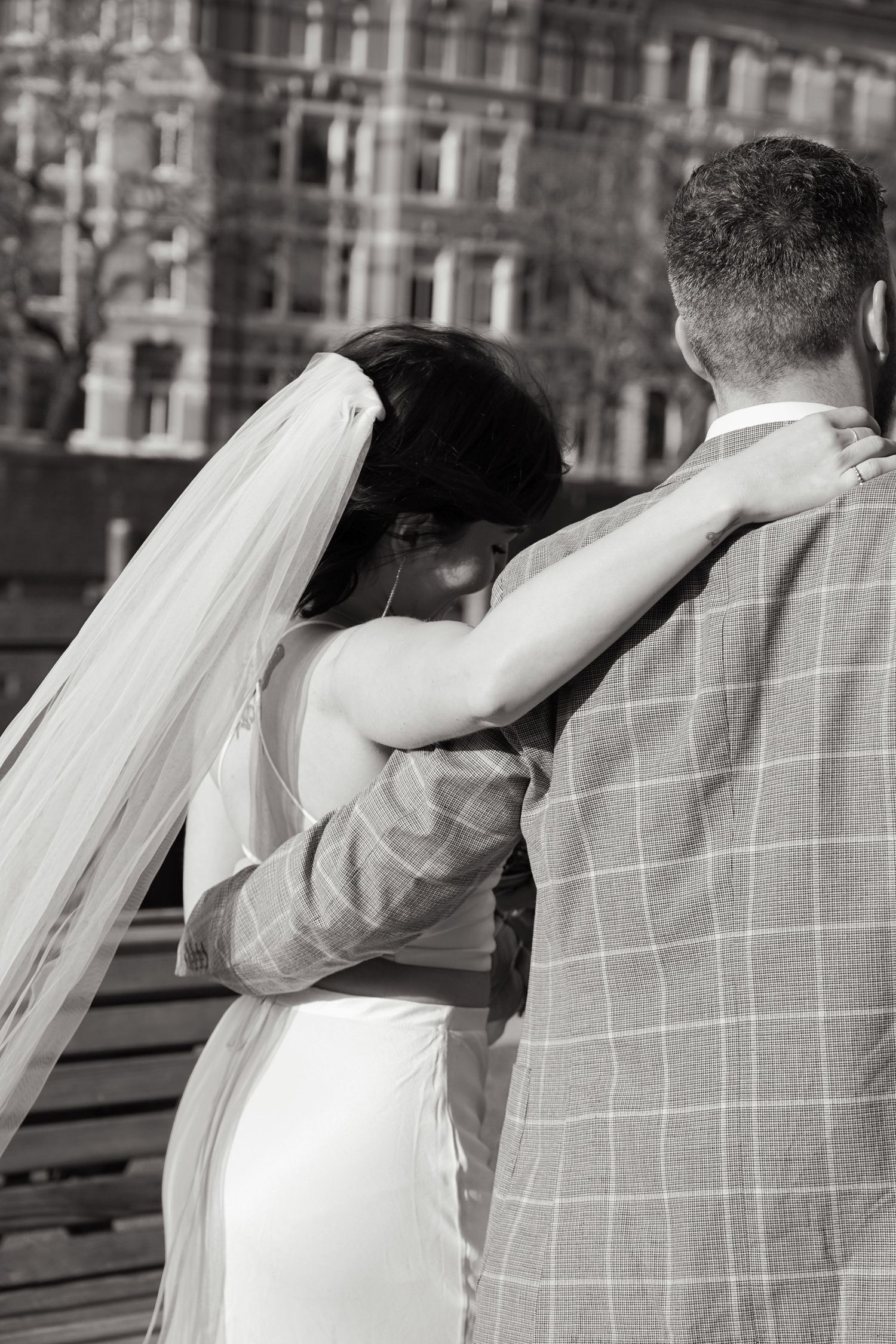 Black and white portrait of couple embracing on deck with urban architecture behind them.