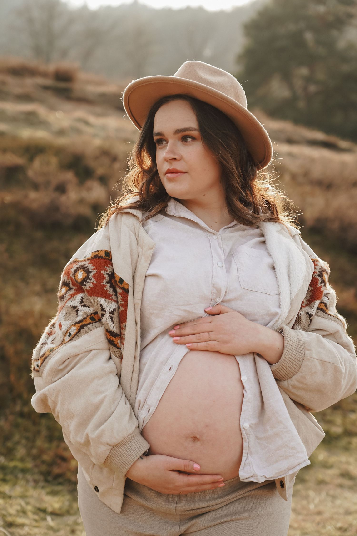 A maternity photo showcasing a tan hat and patterned jacket against a rustic outdoor landscape.