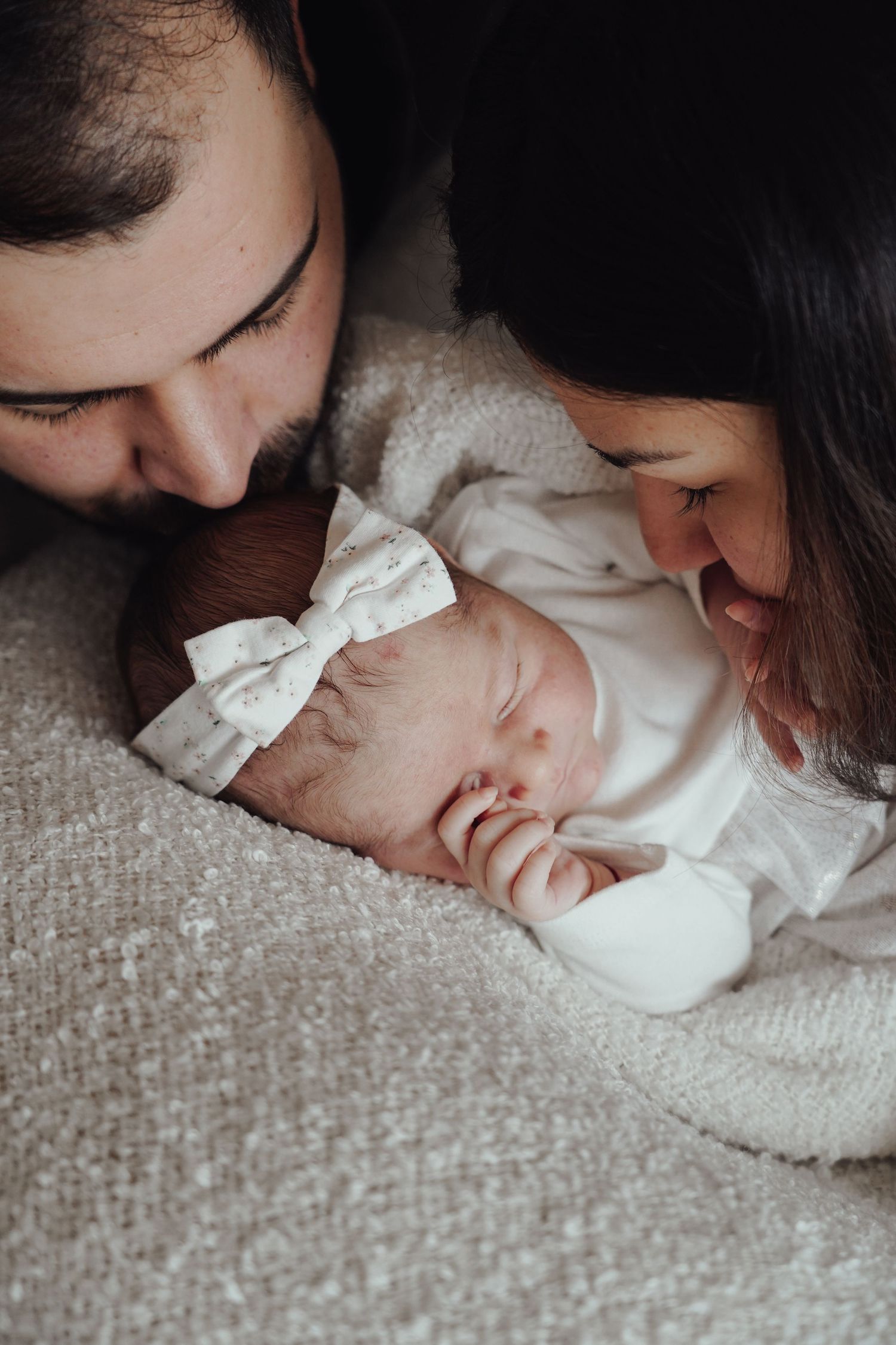 A sleeping newborn baby wearing a white outfit with a bow rests between their parents.