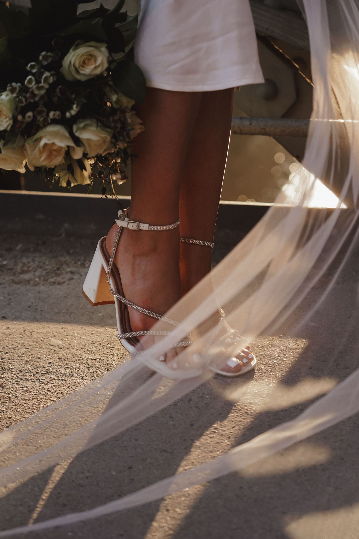 Close-up of bridal shoes and flowing veil with flower bouquet in soft lighting.