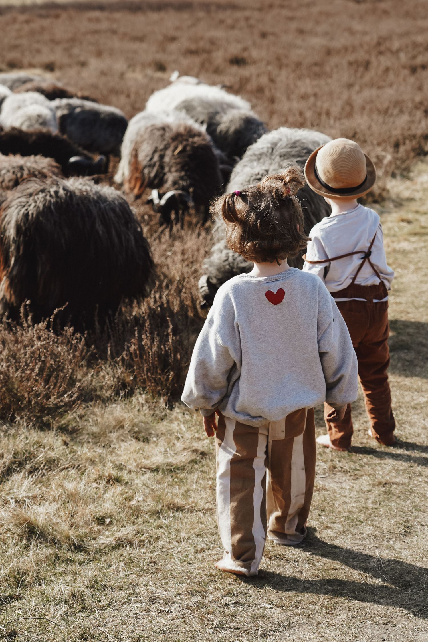 Children in rustic clothing walk near a flock of sheep in a pastoral setting.