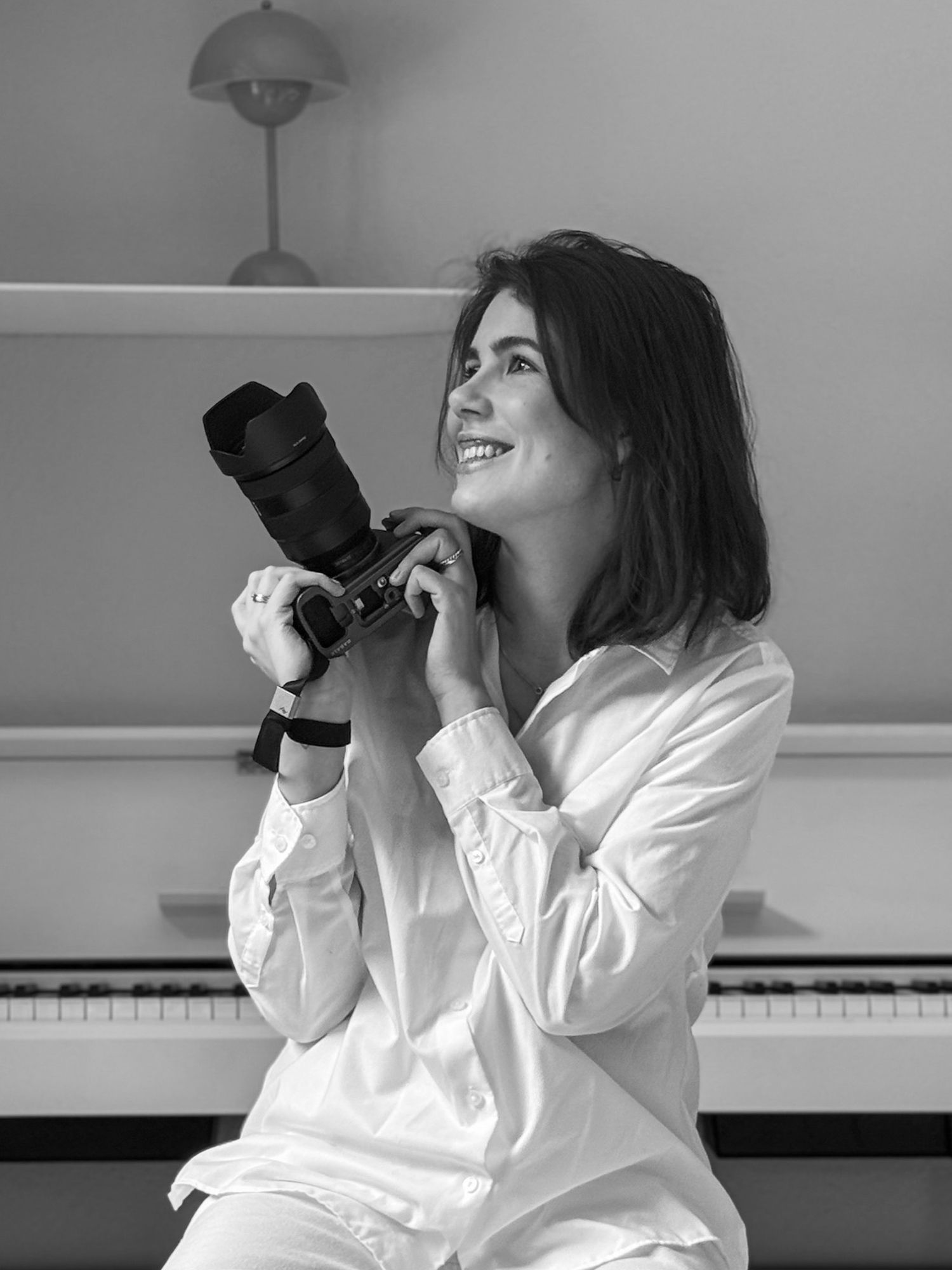 A photographer in white clothing smiles while holding a camera indoors.