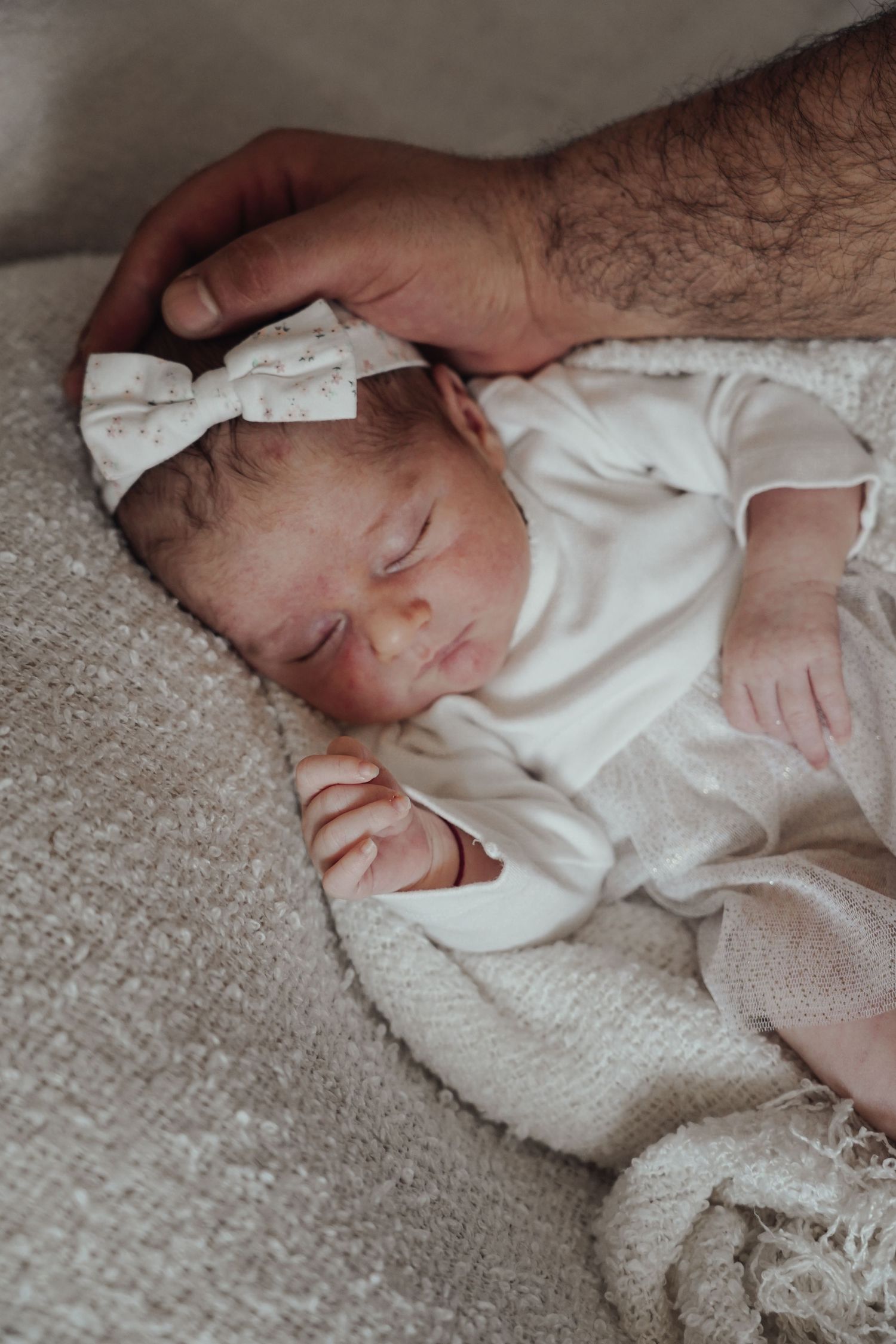 Sleeping newborn in white outfit with bow headband rests on textured gray fabric.