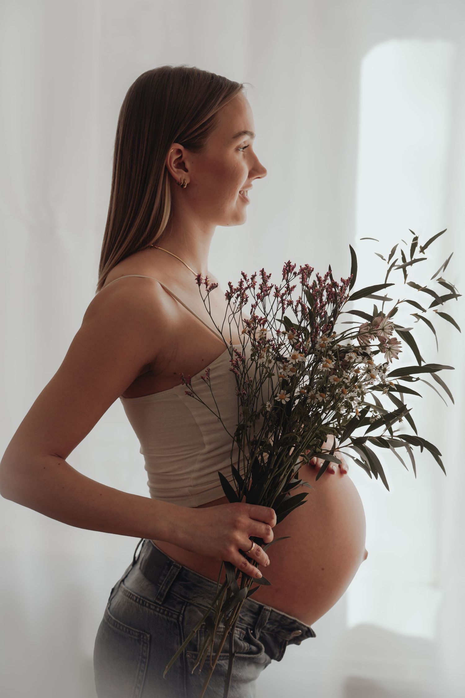 Side profile of an expectant mother in jeans and tank top holding dried flowers against a bright window.